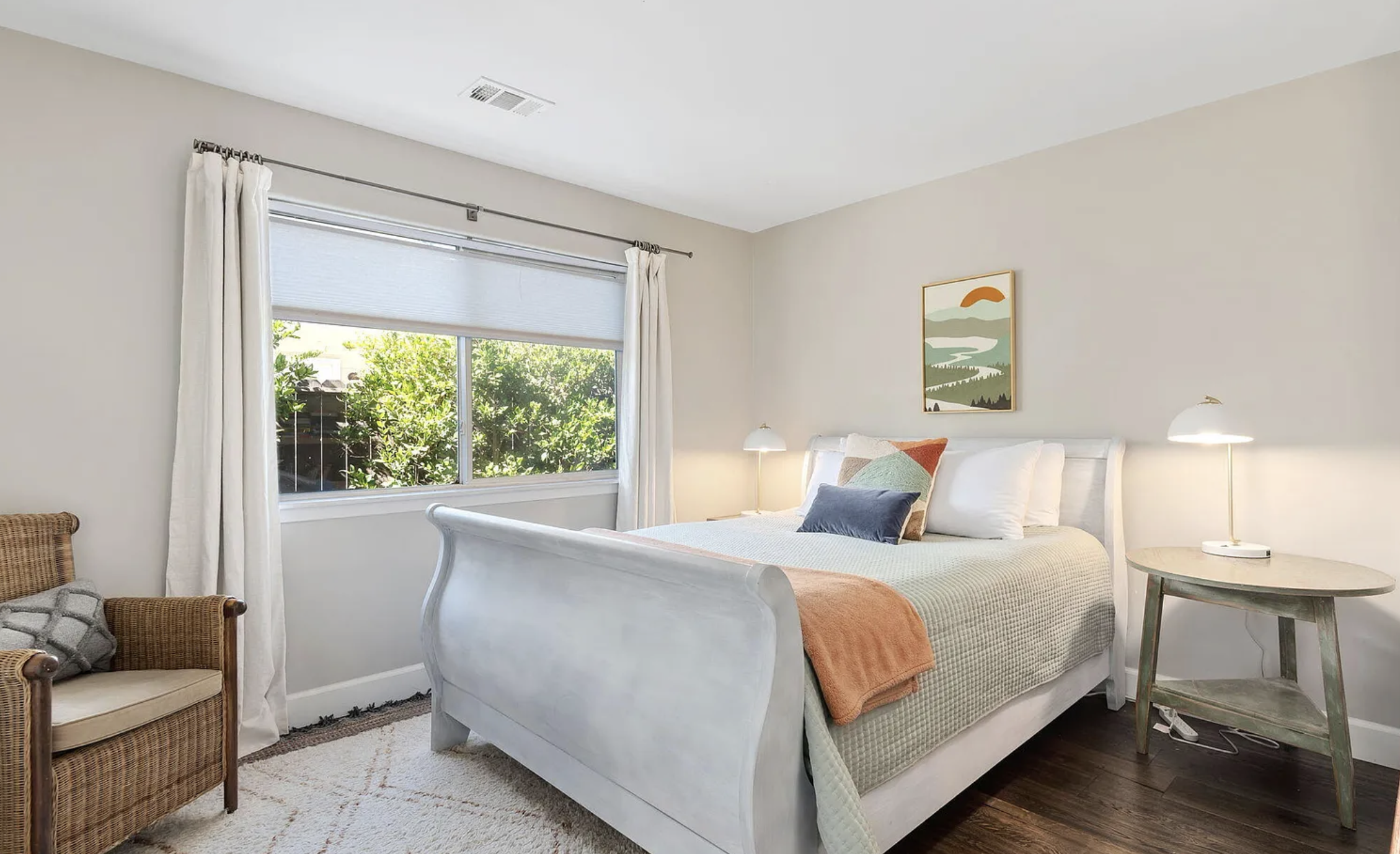 A cozy bedroom with a white bed, colorful pillows, a side table with a lamp, a window with curtains, and a piece of wall art above the bed.