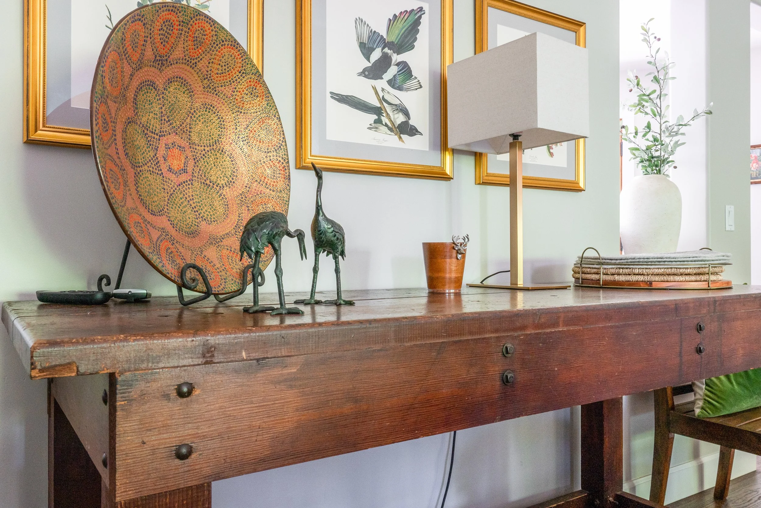 A rustic wooden console table decorated with a large colorful circular art piece, two brass crane sculptures, a small deer figurine, a white lamp with a beige shade, a potted plant, and some neatly stacked books or magazines. Framed bird artwork hang