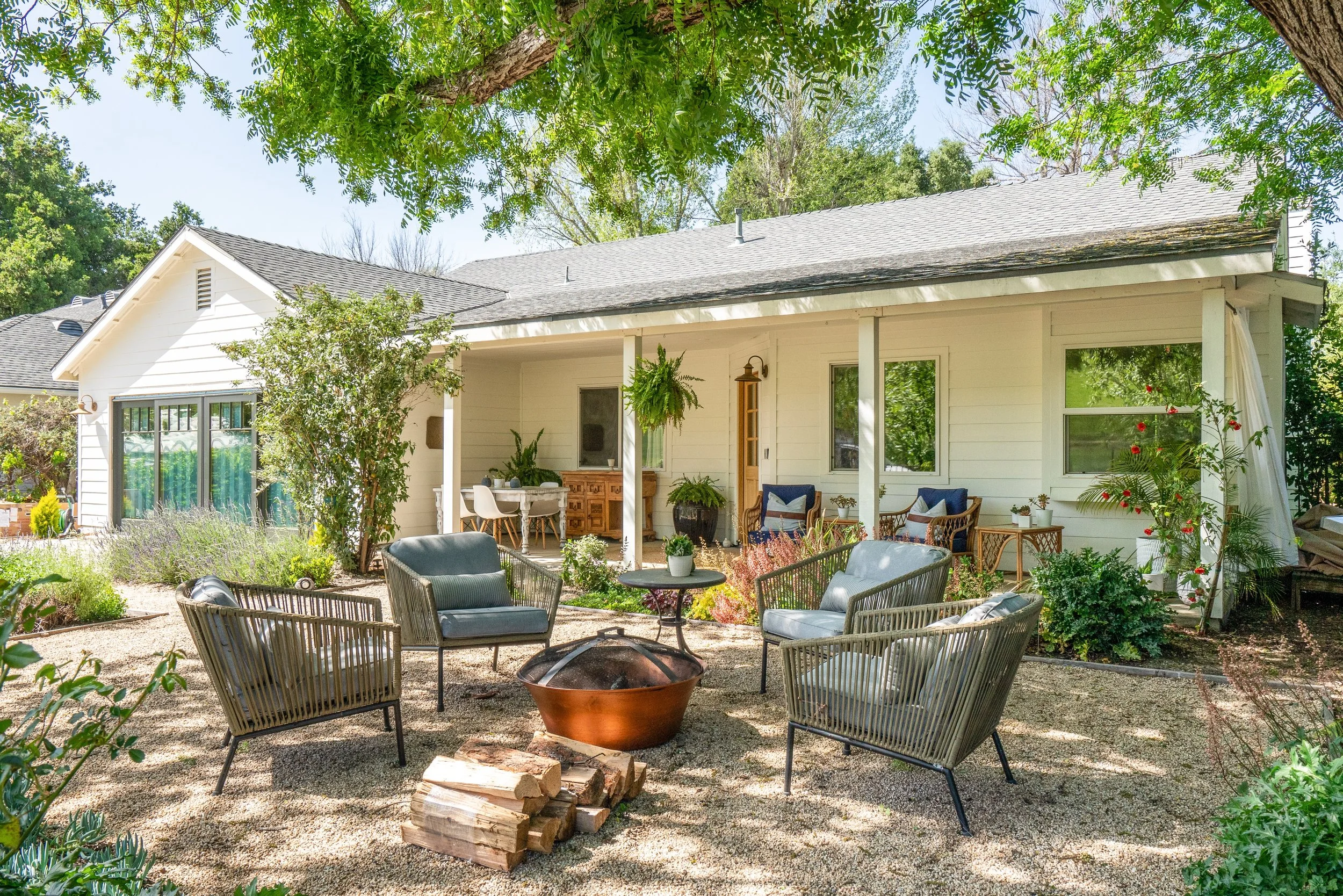 A backyard patio with four modern outdoor chairs surrounding a fire pit, with a stack of firewood nearby. There is a house with a porch in the background decorated with various plants and outdoor furniture, shaded by trees.