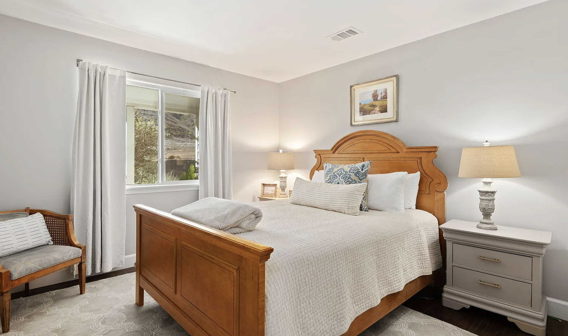 Bedroom with a wooden bed, nightstands, lamps, framed wall art, window with white curtains, beige bedding, and a small upholstered bench.