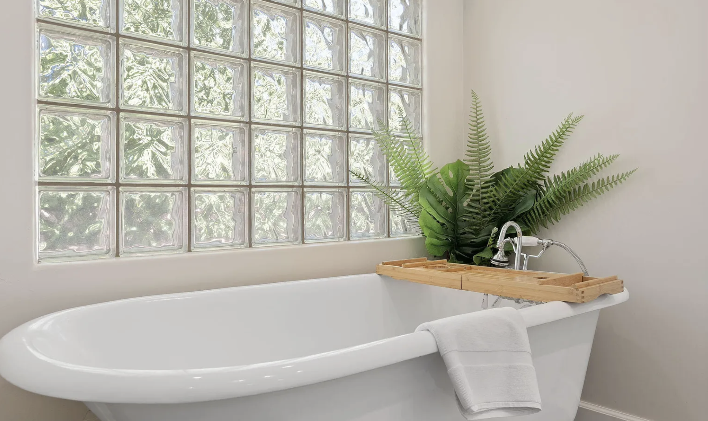 A white bathtub with a wooden bath tray holding a black soap dispenser and a small wooden box, with a white towel draped over the edge, positioned in front of a frosted glass block window with a green indoor plant located behind the tub.