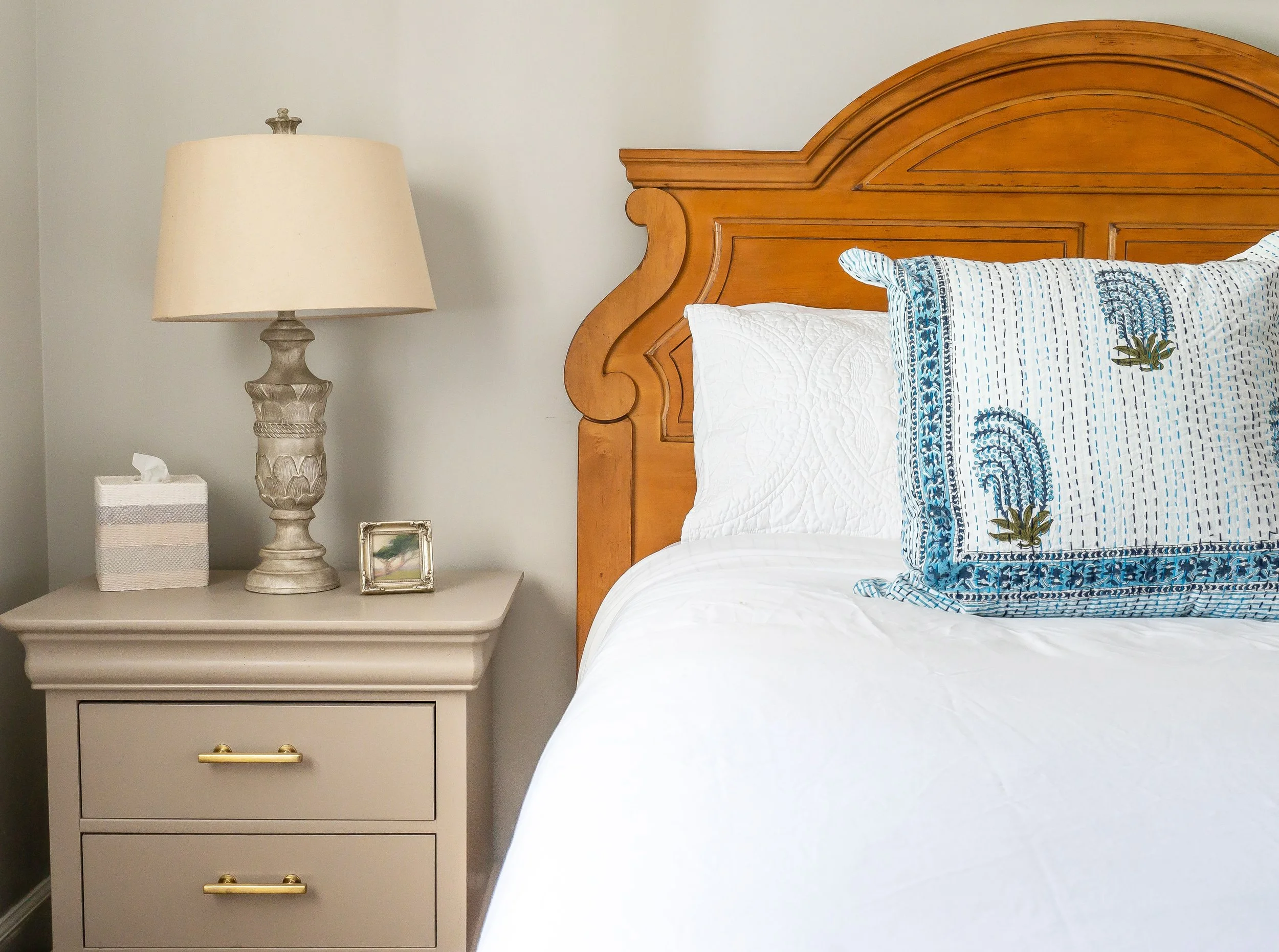 A bedroom corner with a wooden headboard, a white quilt, a pillow with blue embroidery, a beige nightstand, a beige lamp, a small framed picture, and a tissue box.