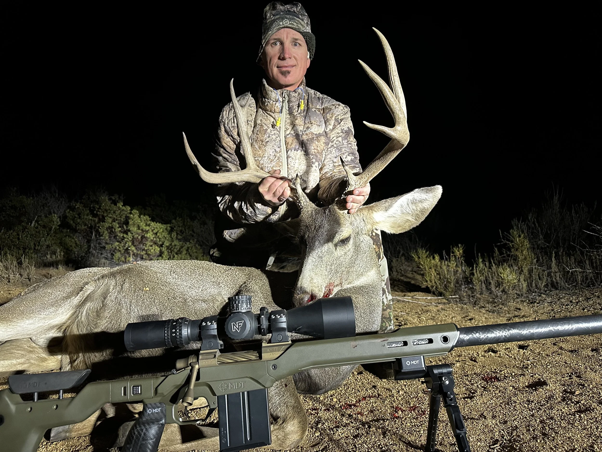 A man in camouflage clothing holding antlers next to a large dead deer with a rifle and scope in the foreground, outdoors at night.