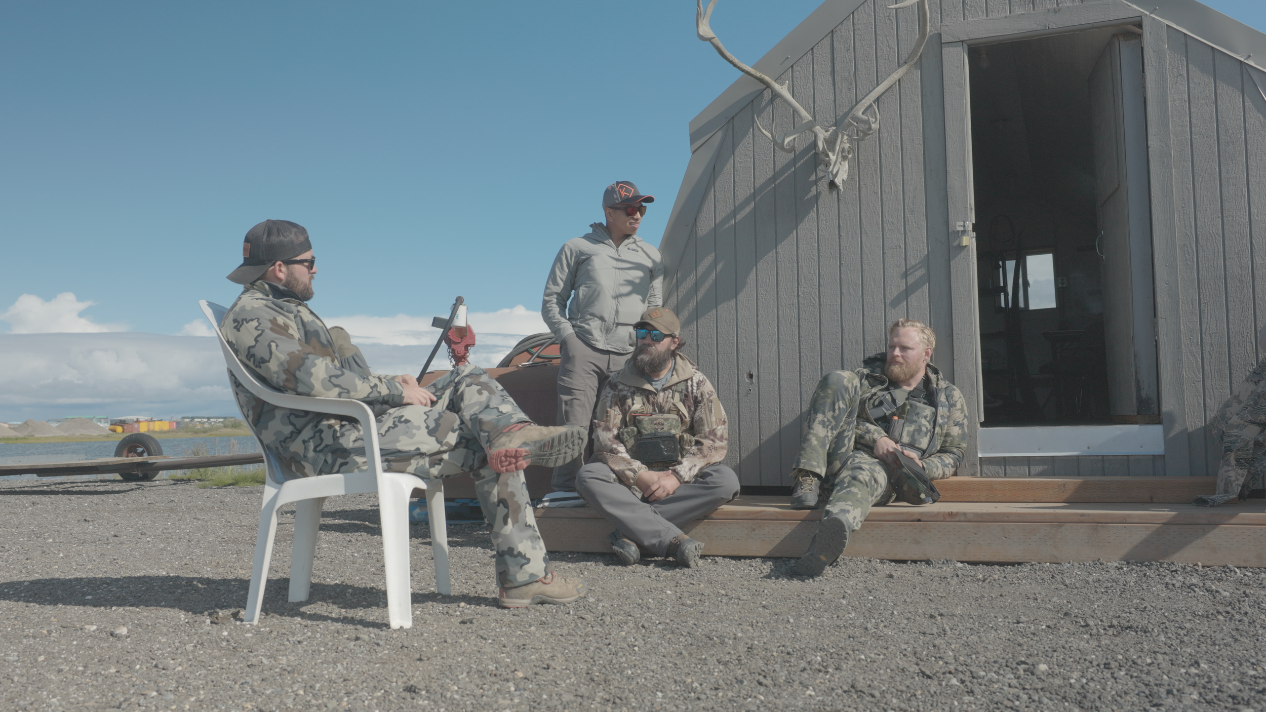 Group of four men, three in camouflage and one in gray, sitting and standing outside a gray shack with a clear blue sky in the background. They are wearing outdoor gear and sunglasses, with one holding a rifle.