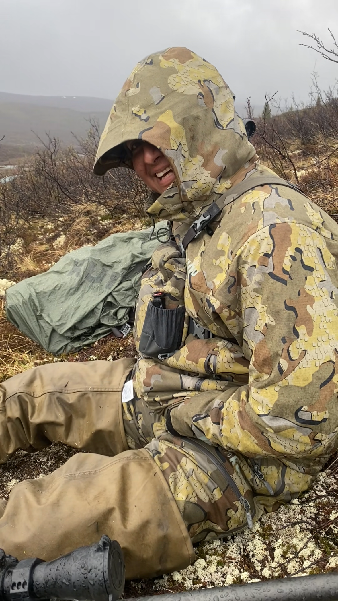 Man in camouflage gear sitting outdoors on a cloudy day, surrounded by sparse bushes and low vegetation.