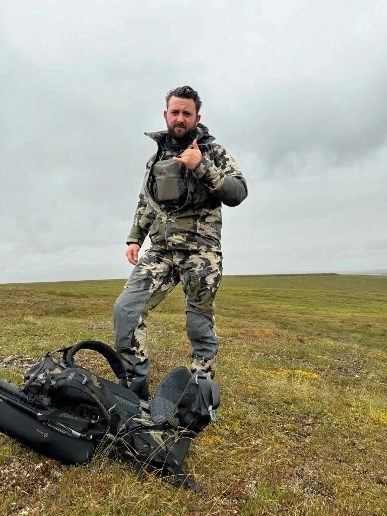 Man in camouflage clothing standing outdoors on grassy terrain under cloudy sky, giving a thumbs-up gesture, with a backpack on the ground nearby.