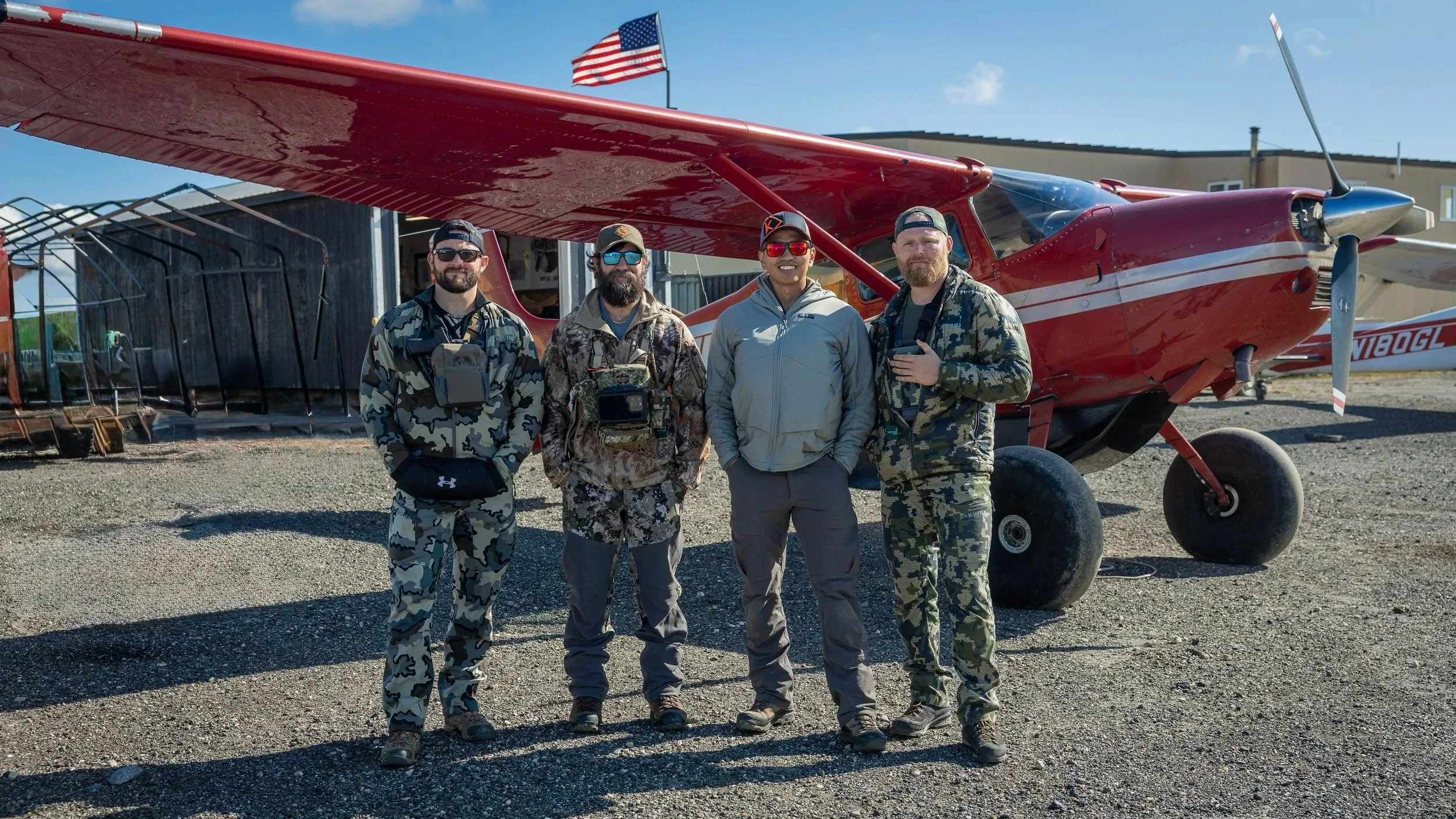Four men standing in front of a red small airplane with an American flag on top. They are dressed in camouflage and outdoor gear, smiling at the camera under a clear blue sky.