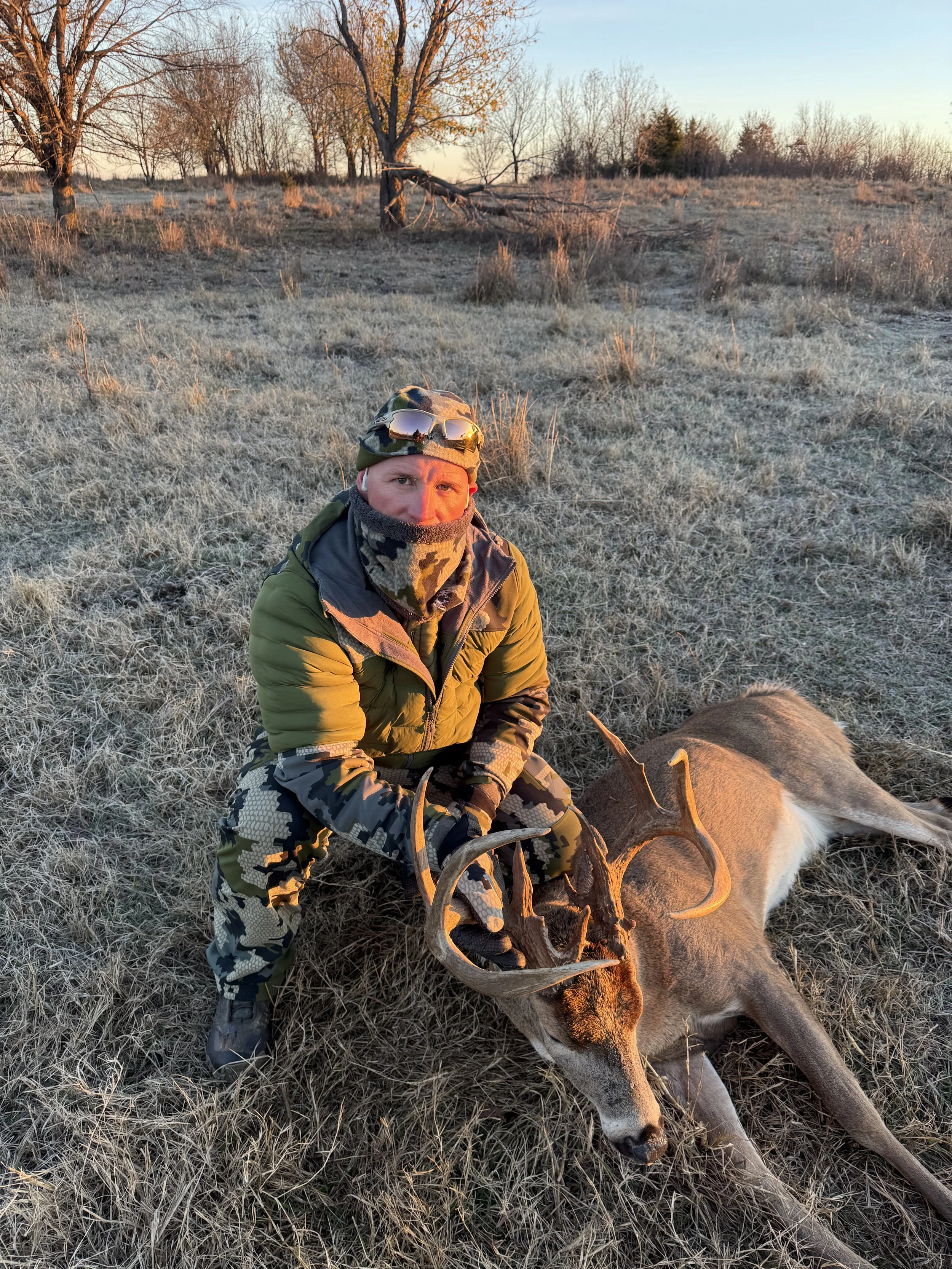 A man dressed in camouflage and outdoor gear kneels on frosty grass, holding the antlers of a large deer with a buck's antlers, lying on the ground. The background features leafless trees and clear skies at sunset or sunrise.
