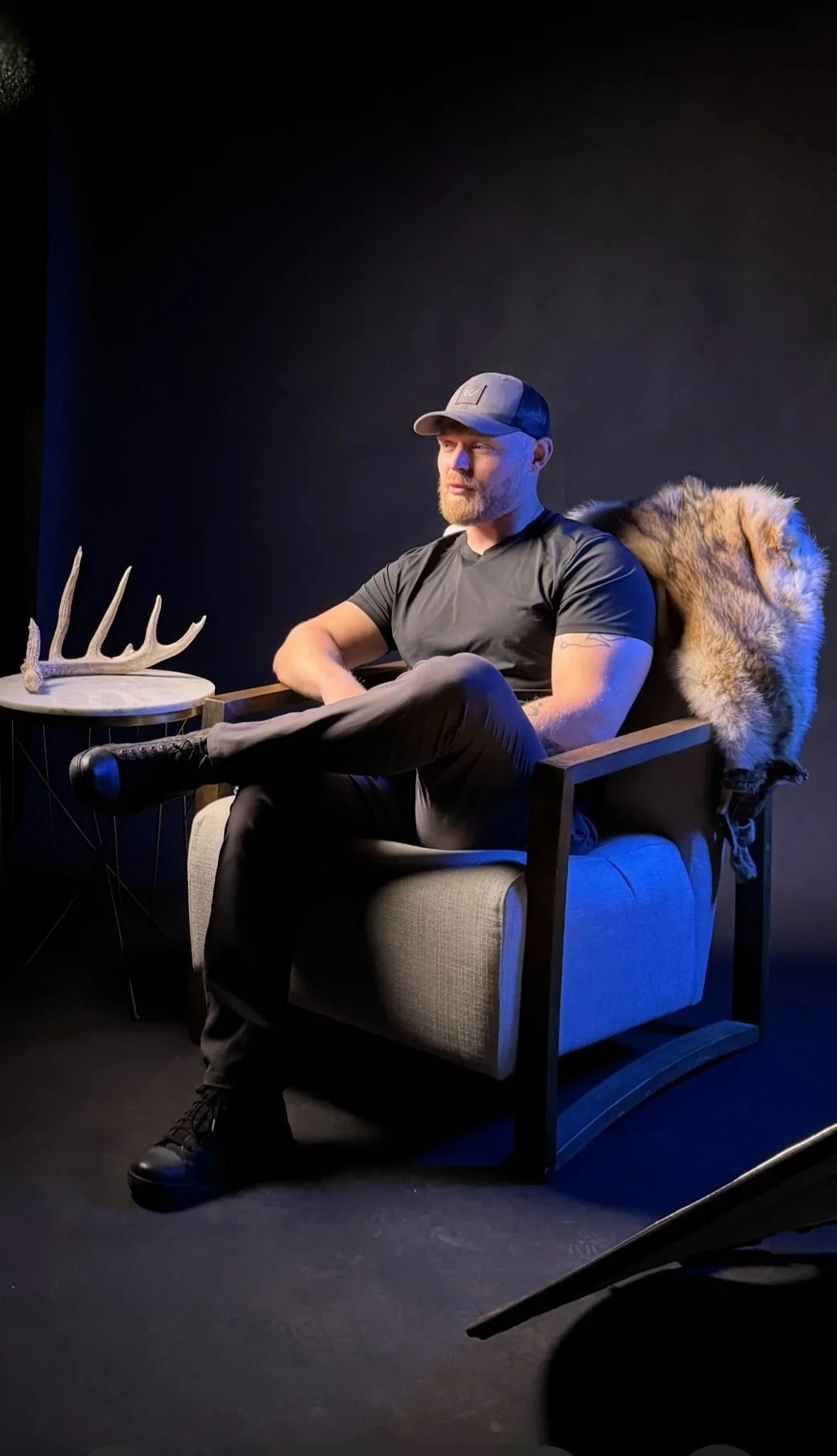 A man with a beard and baseball cap sitting in a modern armchair with a wolf fur on the backrest, against a dark background, with a side table holding deer antlers next to him.