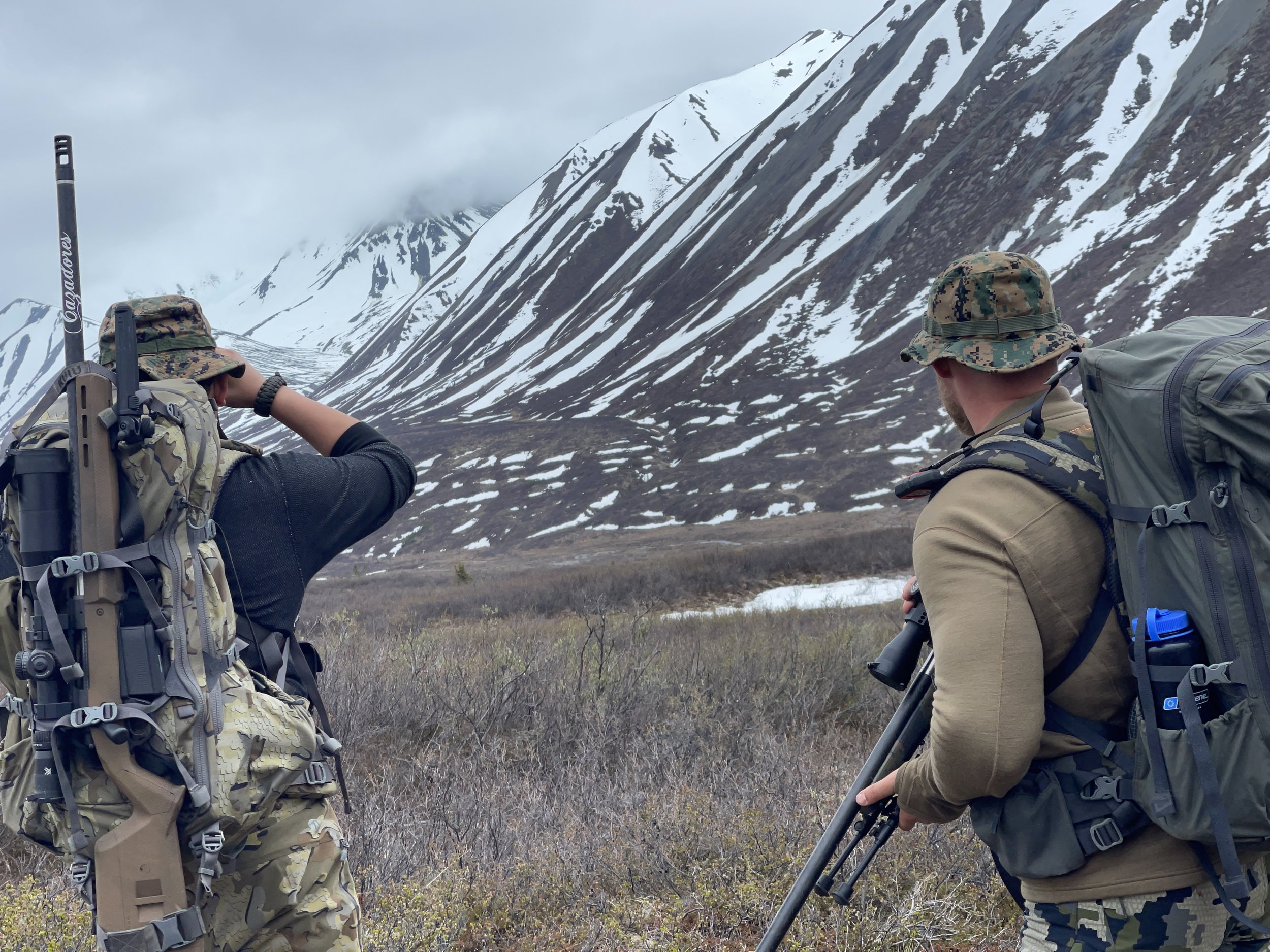 Two men hiking in a mountainous area with snow patches, wearing camouflage and outdoor gear, one using binoculars and the other with a long object, possibly a rifle or camera, on their backpacks.