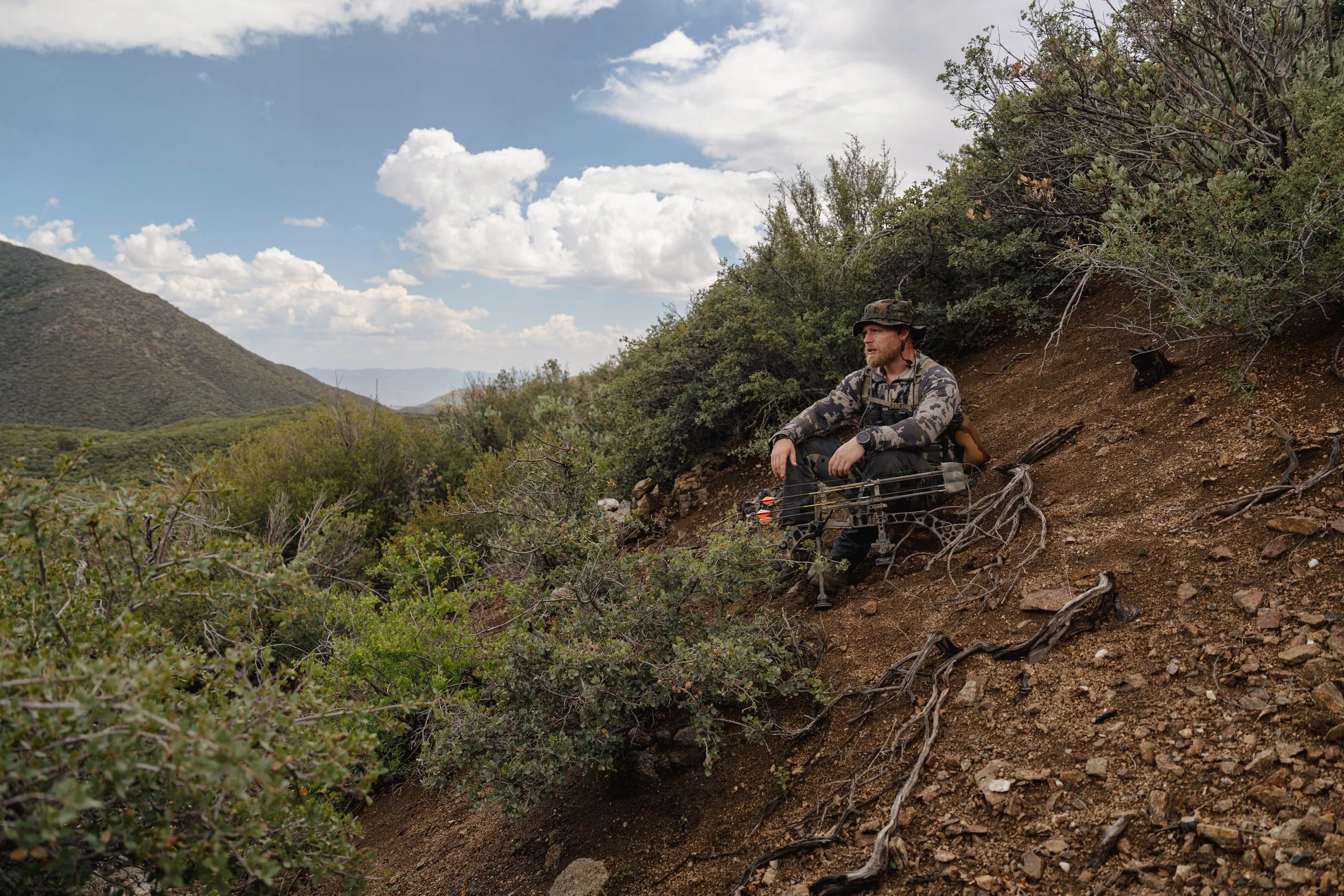 A man sitting on a hillside with vegetation and hills in the background, wearing camouflage clothing and a hat, with a prosthetic leg.