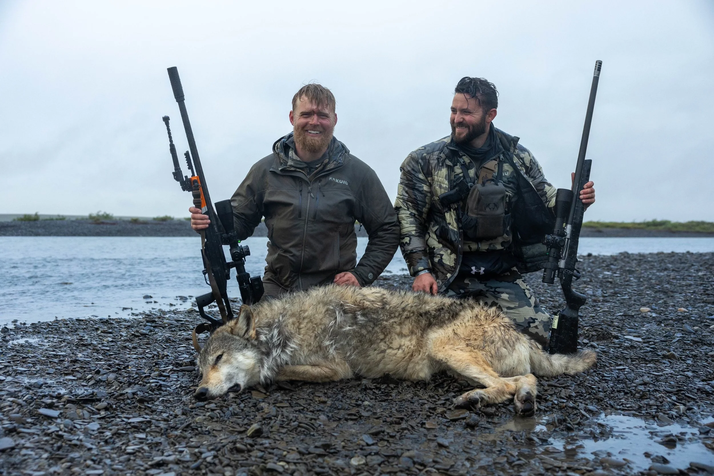 Two men with rifles kneeling on rocky shore next to a lying wolf near a body of water.