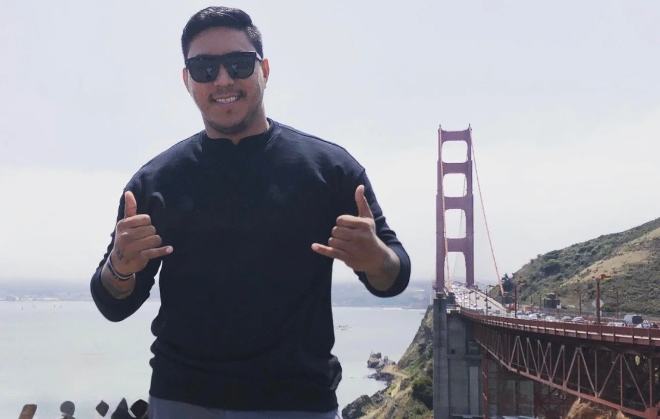 Smiling man wearing sunglasses making shaka hand gestures in front of the Golden Gate Bridge in San Francisco.