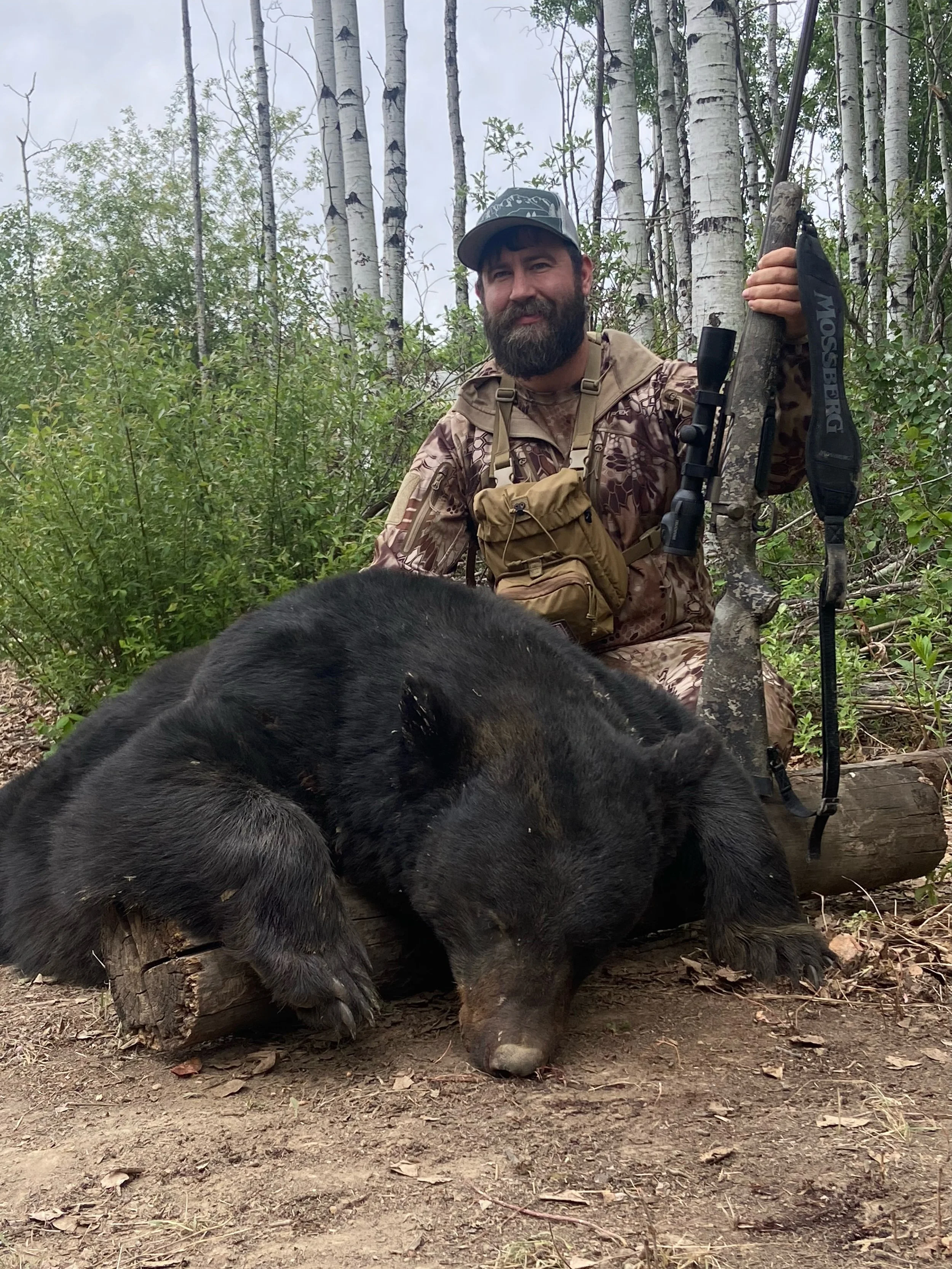 A man in camouflage hunting gear, smiling, kneeling in a forest beside a large, dead black bear that is lying on the ground.