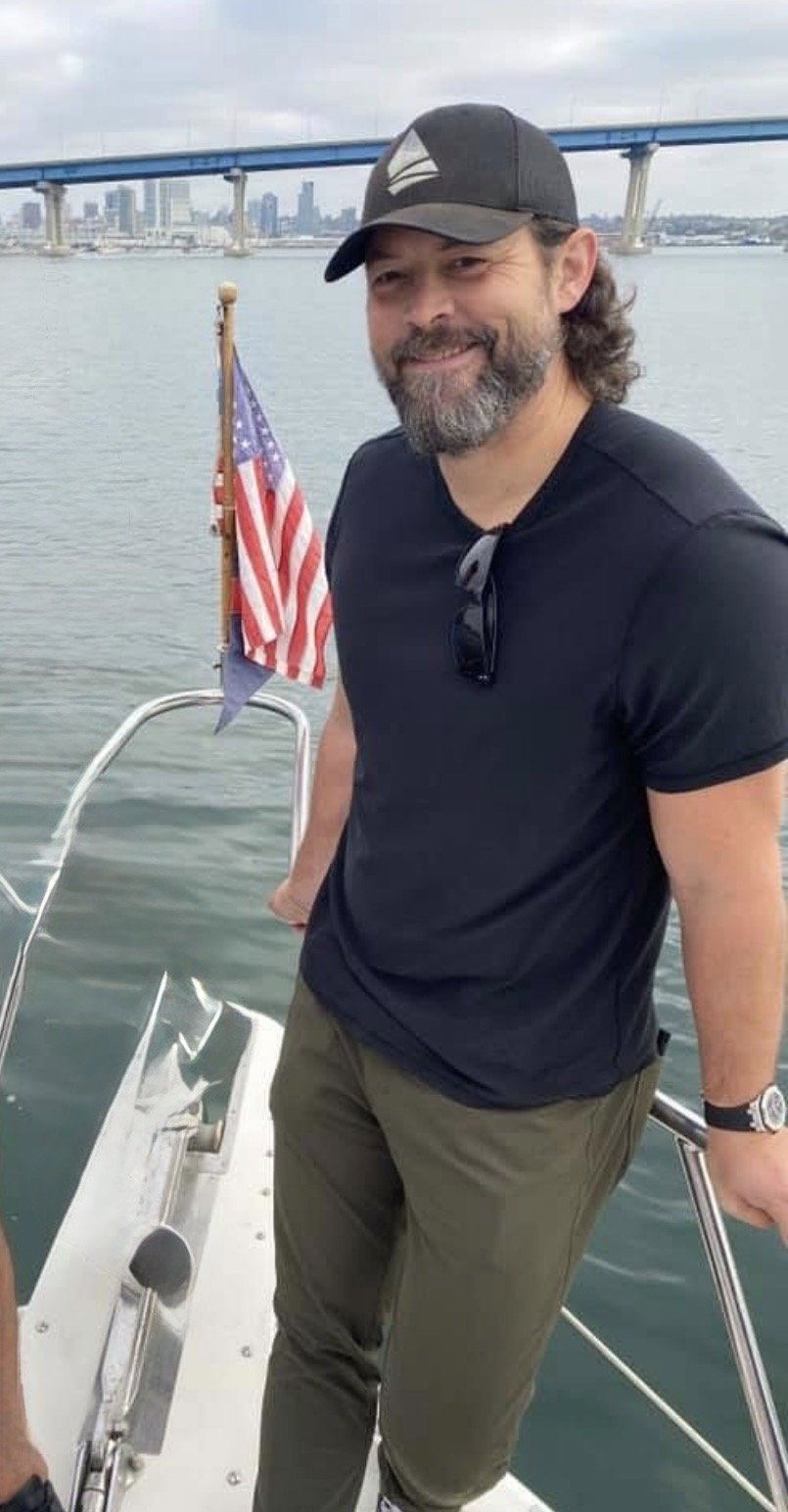 A man with a beard and long hair, wearing a black baseball cap and black t-shirt, stands on a boat in a body of water. An American flag is visible behind him, and a city skyline with a bridge is in the background.