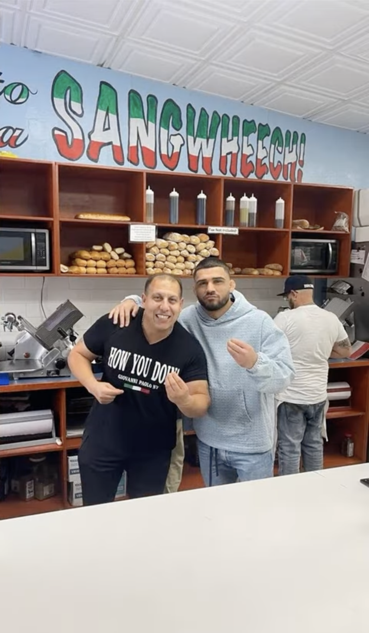 Two men posing with their fists up inside a sandwich shop with shelves of bread and condiments behind them.