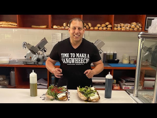 Smiling man wearing a black t-shirt with the words 'Time to Make a Sandwich' written on it, standing behind a counter with two large sandwiches and two bottles of condiments, in a kitchen or deli setting.