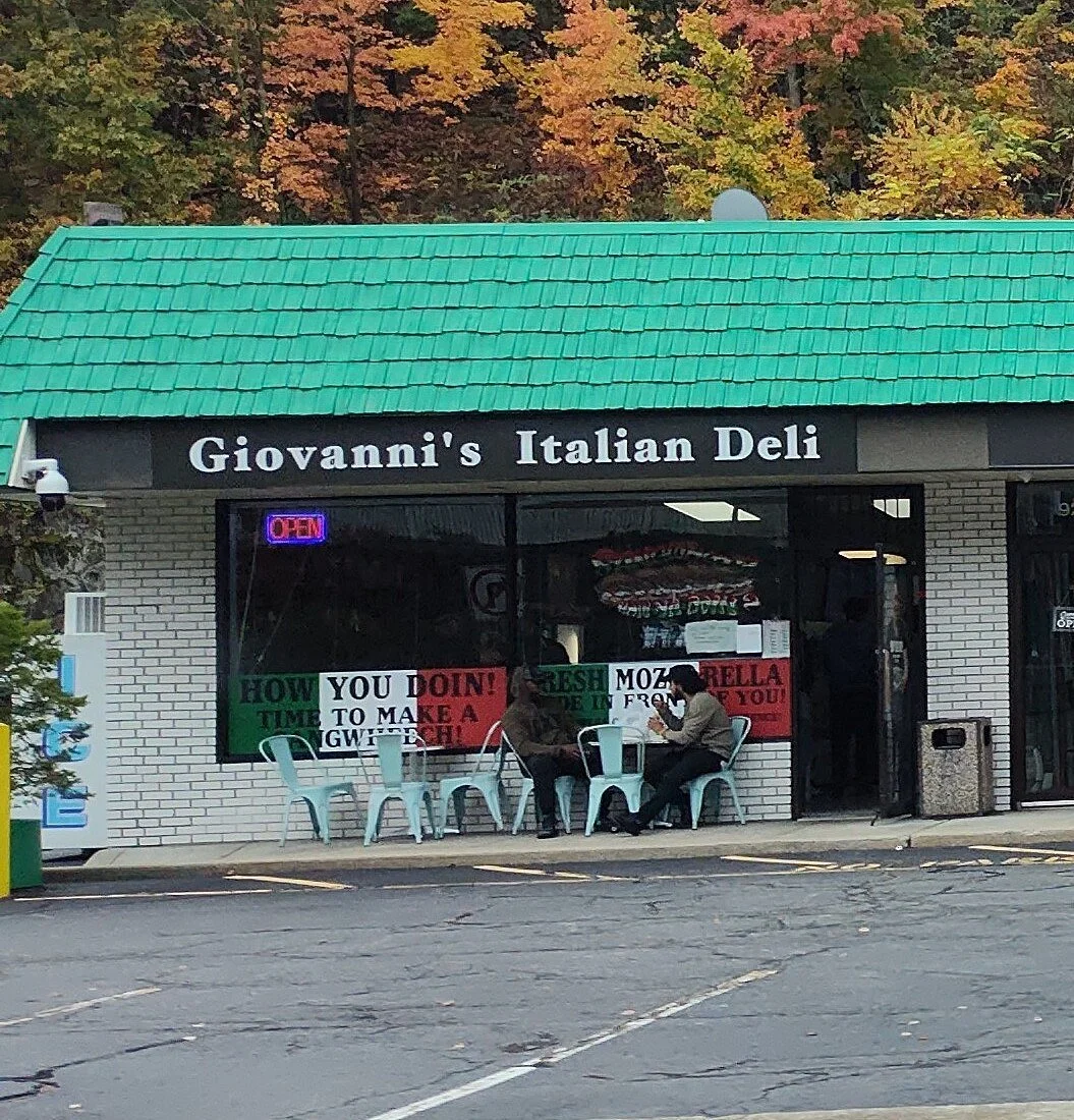 Exterior view of Giovanni's Italian Deli with a green roof and a black sign. Three people sitting outside at a table. Signage includes promotional signs in red, green, and white.