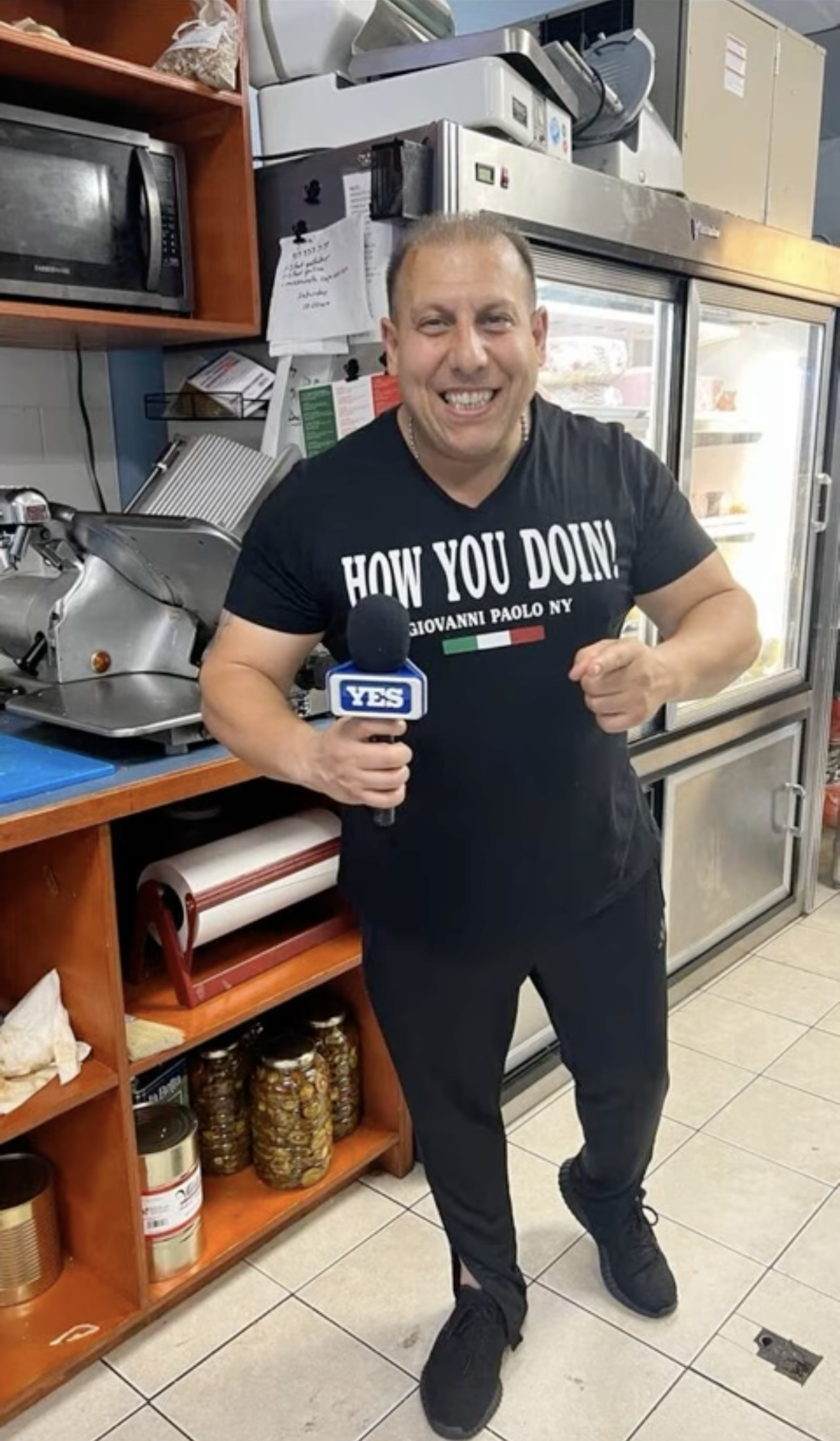 A man in a black T-shirt holding a microphone with the YES logo, standing in a commercial kitchen, smiling and pointing at himself. Behind him are kitchen appliances and shelves with canned goods.