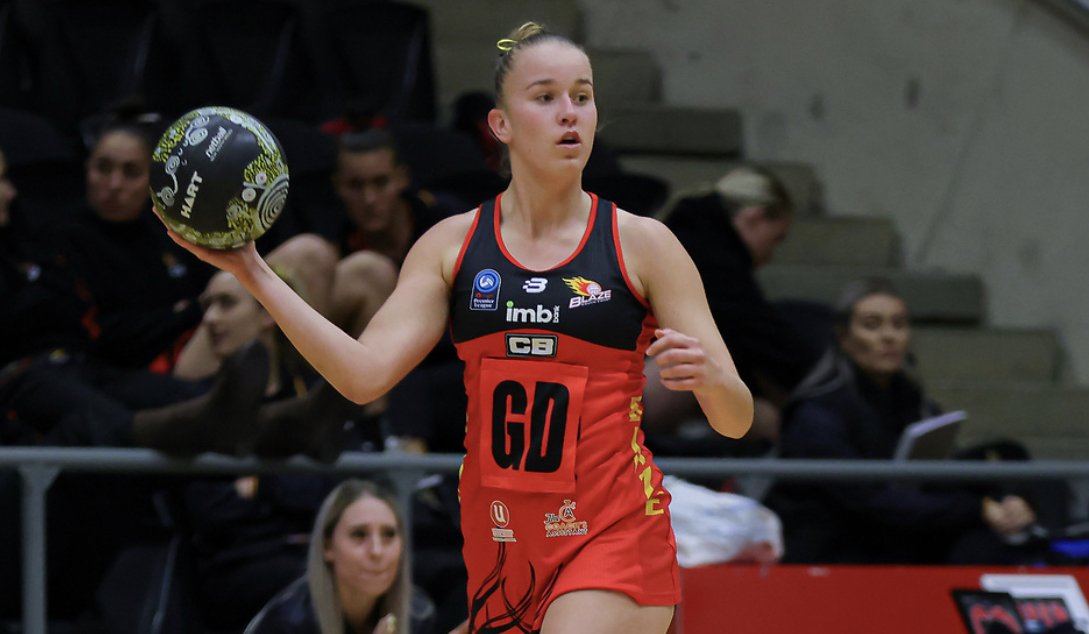A female athlete in a red and black sports uniform holding a volleyball on an indoor court with spectators in the background.