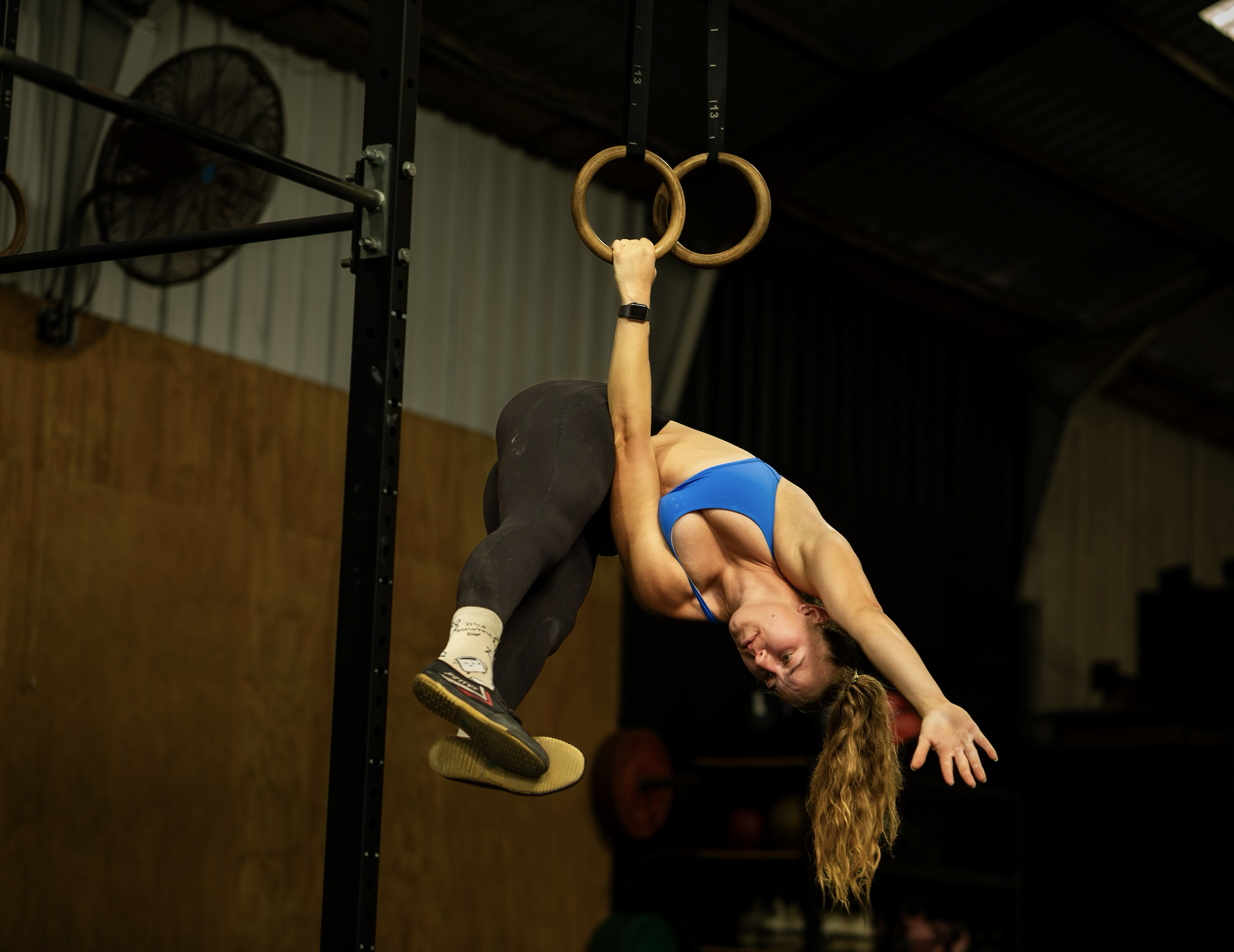 Female gymnast in blue sports bra and black leggings hanging upside down on gymnastic rings in a gym.