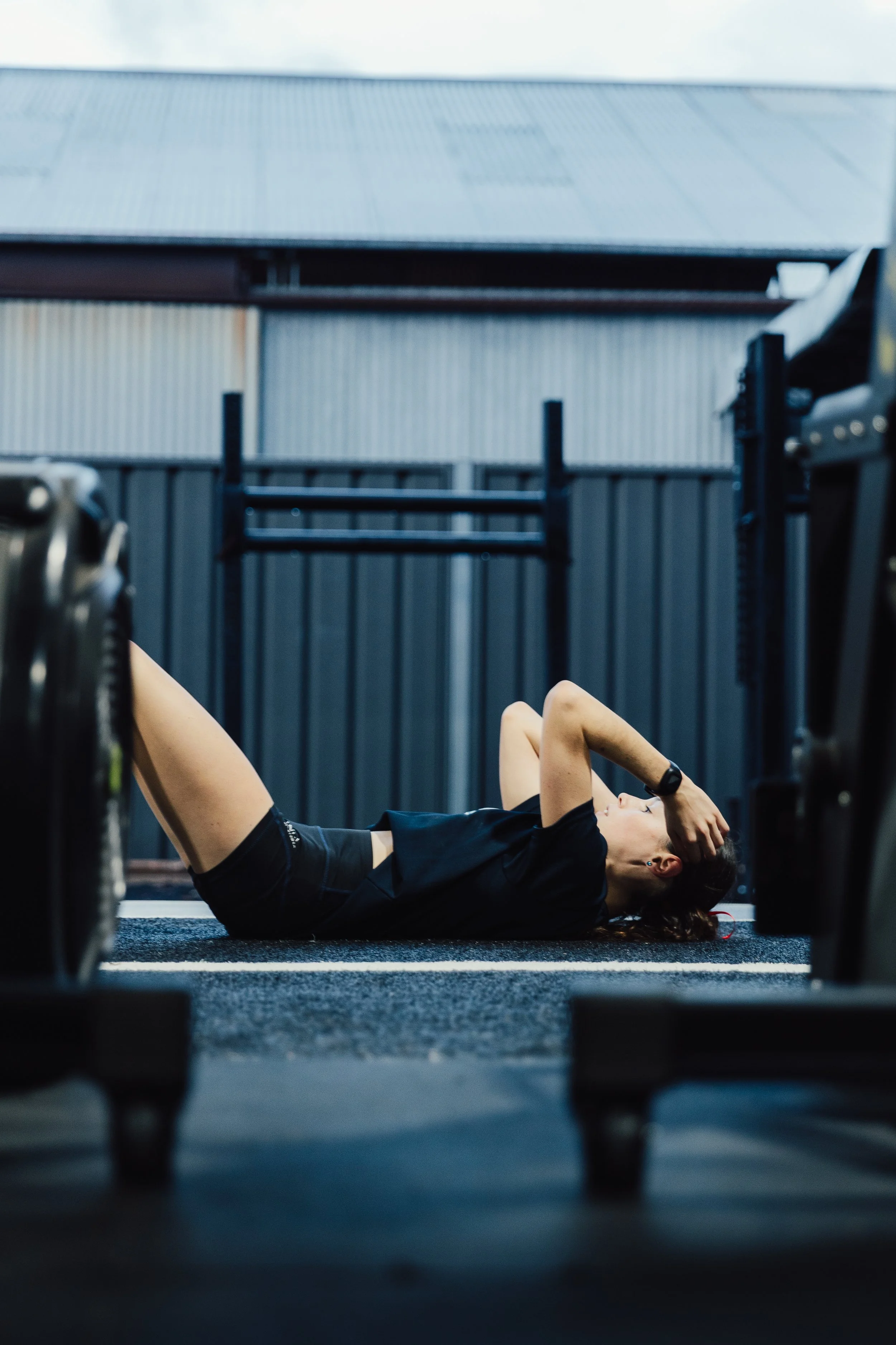 A woman lying on her back on a gym floor, resting after a workout, with a black gym outfit, surrounded by workout equipment.