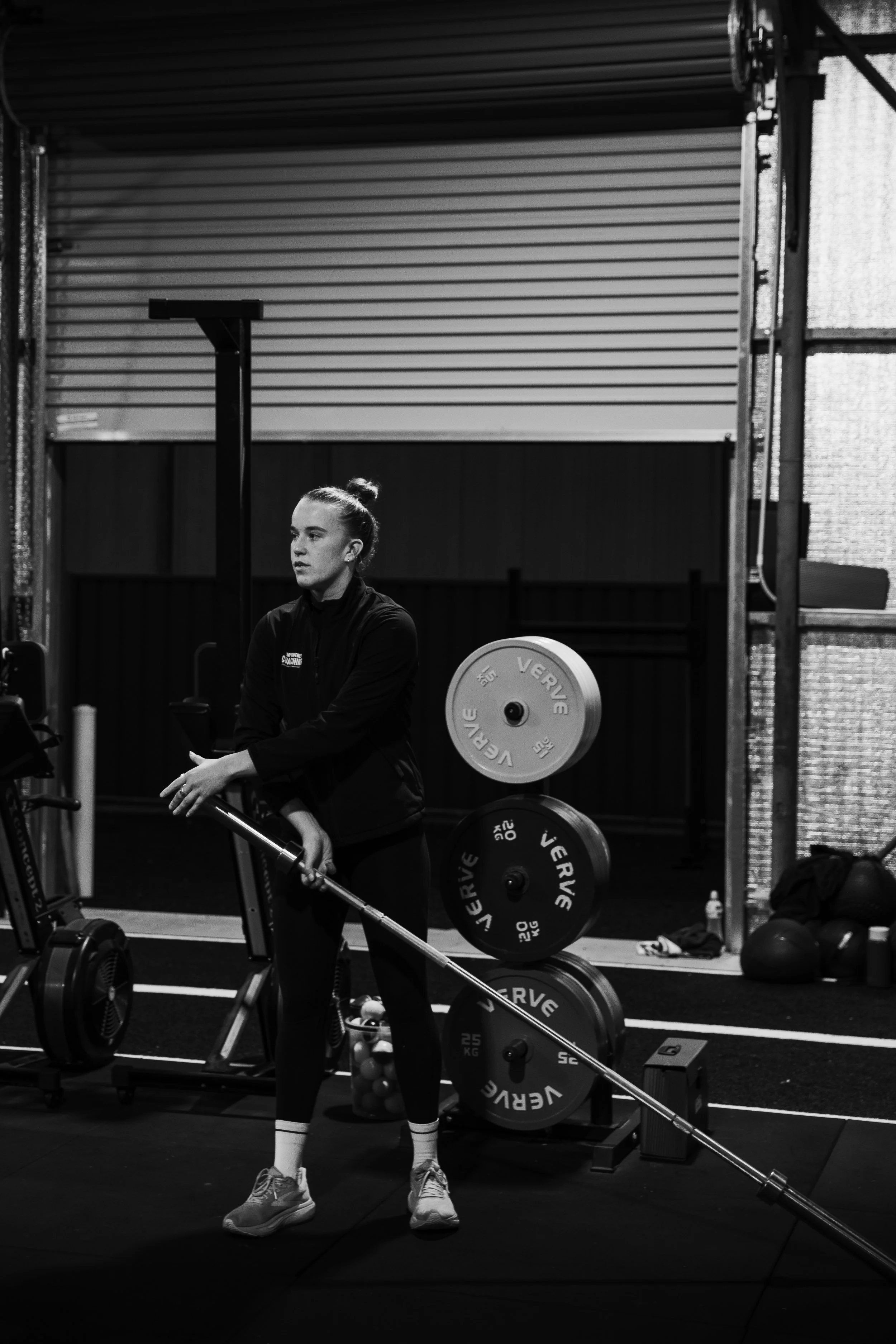 A woman in workout attire in a gym, preparing to lift weights with a barbell, surrounded by gym equipment and weights.