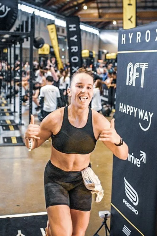 A woman in athletic wear smiling and giving a thumbs-up at a fitness event in a gym with many people in the background.