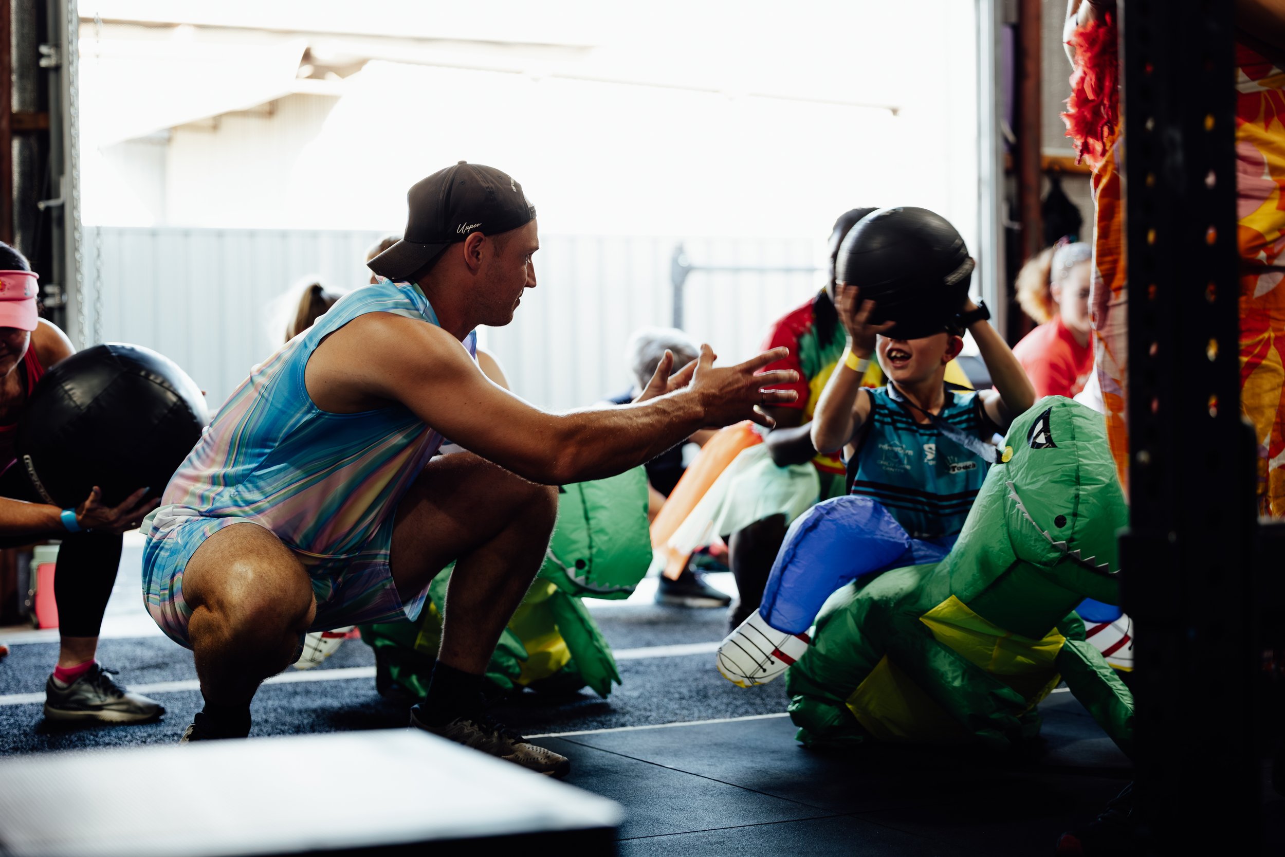 An adult male instructing children during a dinosaur-themed activity at an indoor play area, with children dressed in dinosaur costumes.