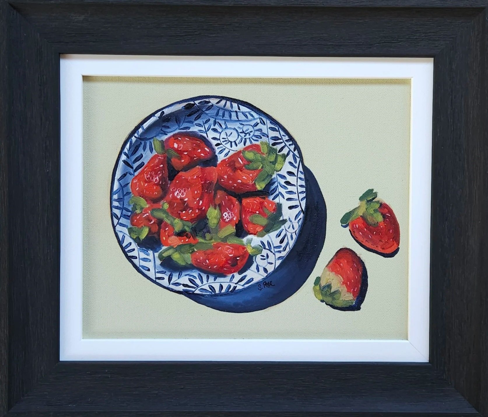 Painting of a bowl of strawberries on a patterned plate with two strawberries outside the bowl, framed in black.
