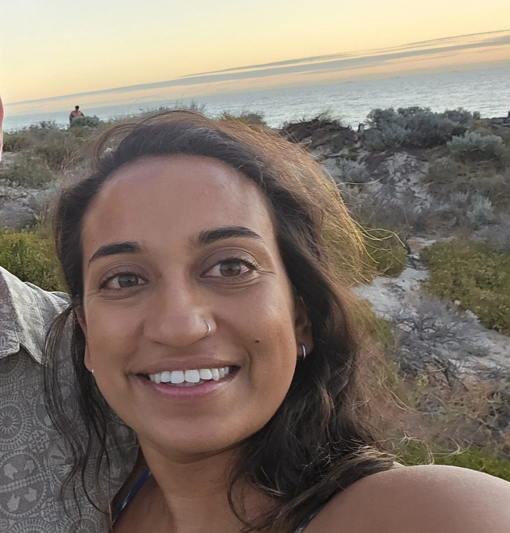 A woman smiling at the camera outdoors with a sunset sky and ocean in the background.