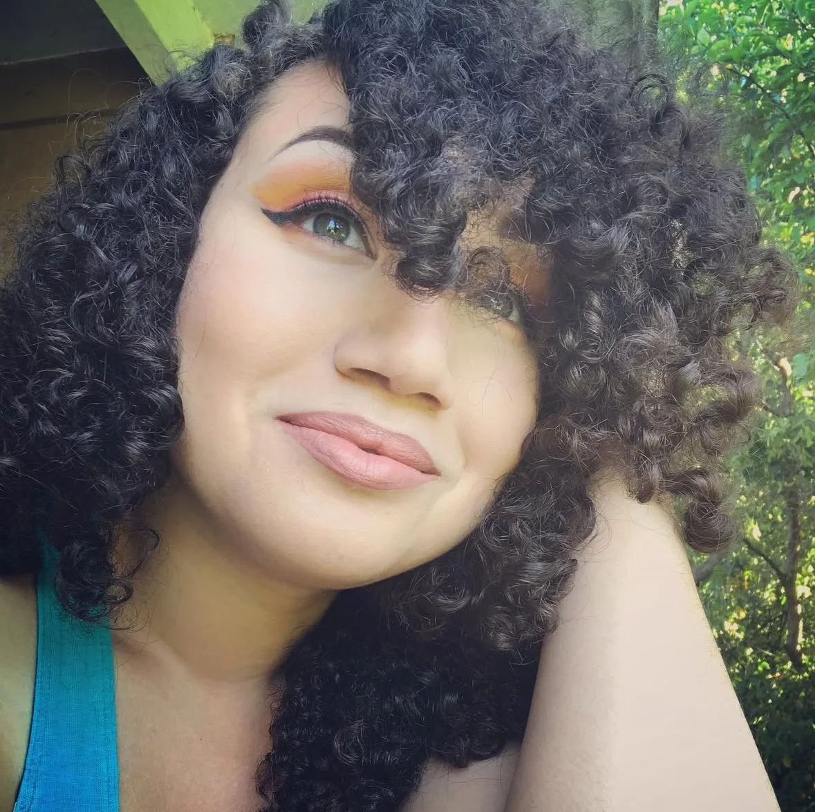 A woman with curly dark hair and vibrant makeup poses outdoors with greenery in the background.