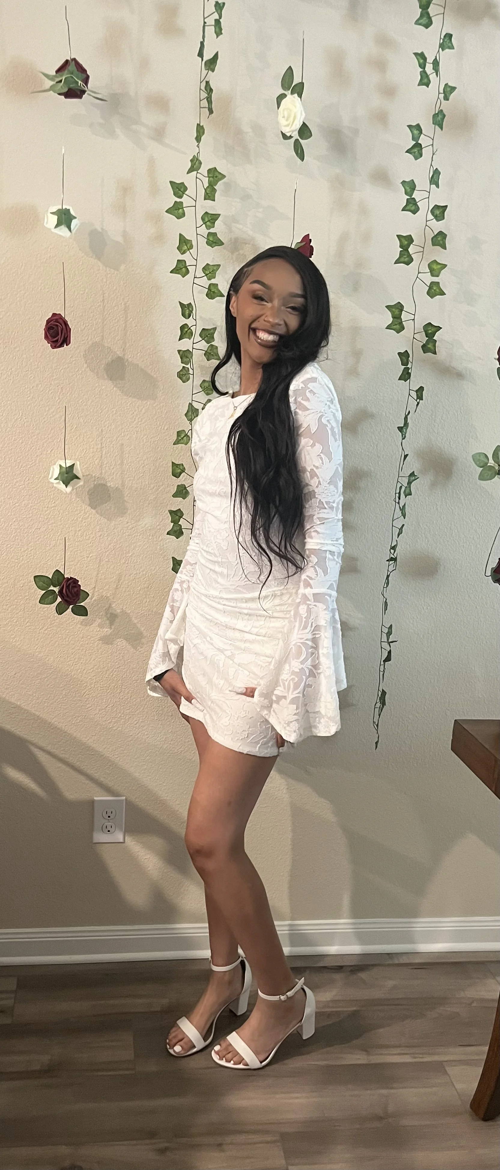 A woman with long black hair smiling in a white lace dress and white high-heeled sandals, standing in front of a wall decorated with hanging roses and ivy.