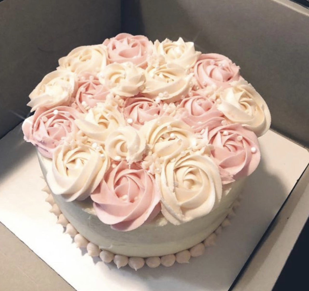 A round cake decorated with pink and white rose-shaped frosting swirls, with small white pearl accents, on a white cake board.