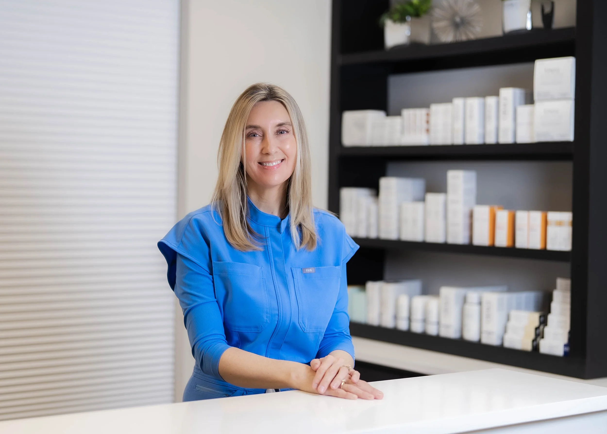 A smiling woman in a blue uniform standing behind a white counter in front of shelves filled with boxes.