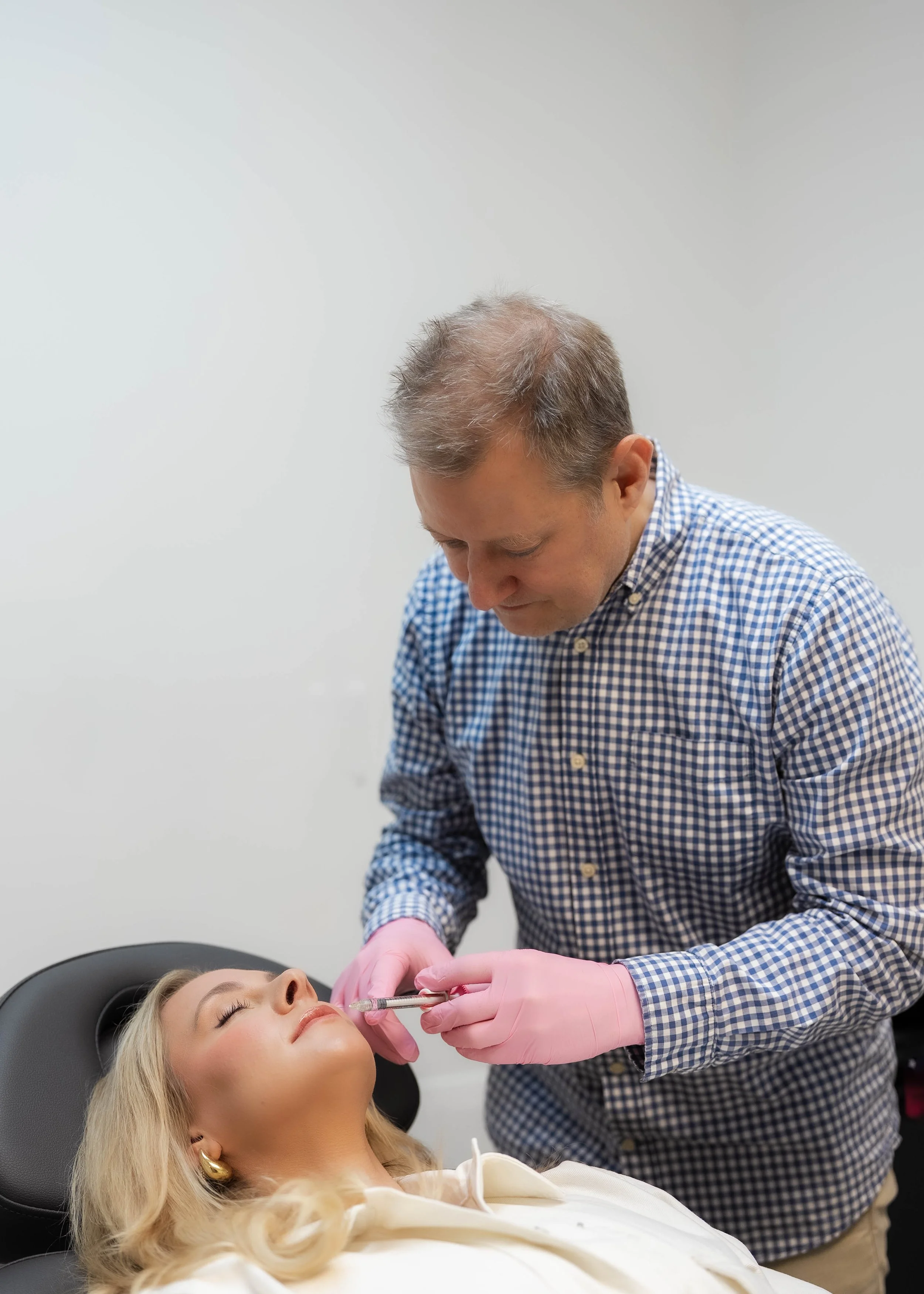 A woman lying back in a dental chair while a man, possibly a dentist, administers a cosmetic facial injection using a syringe. The woman has blonde hair and is wearing makeup, while the man has short, graying hair, and is wearing a checked shirt and pink gloves.