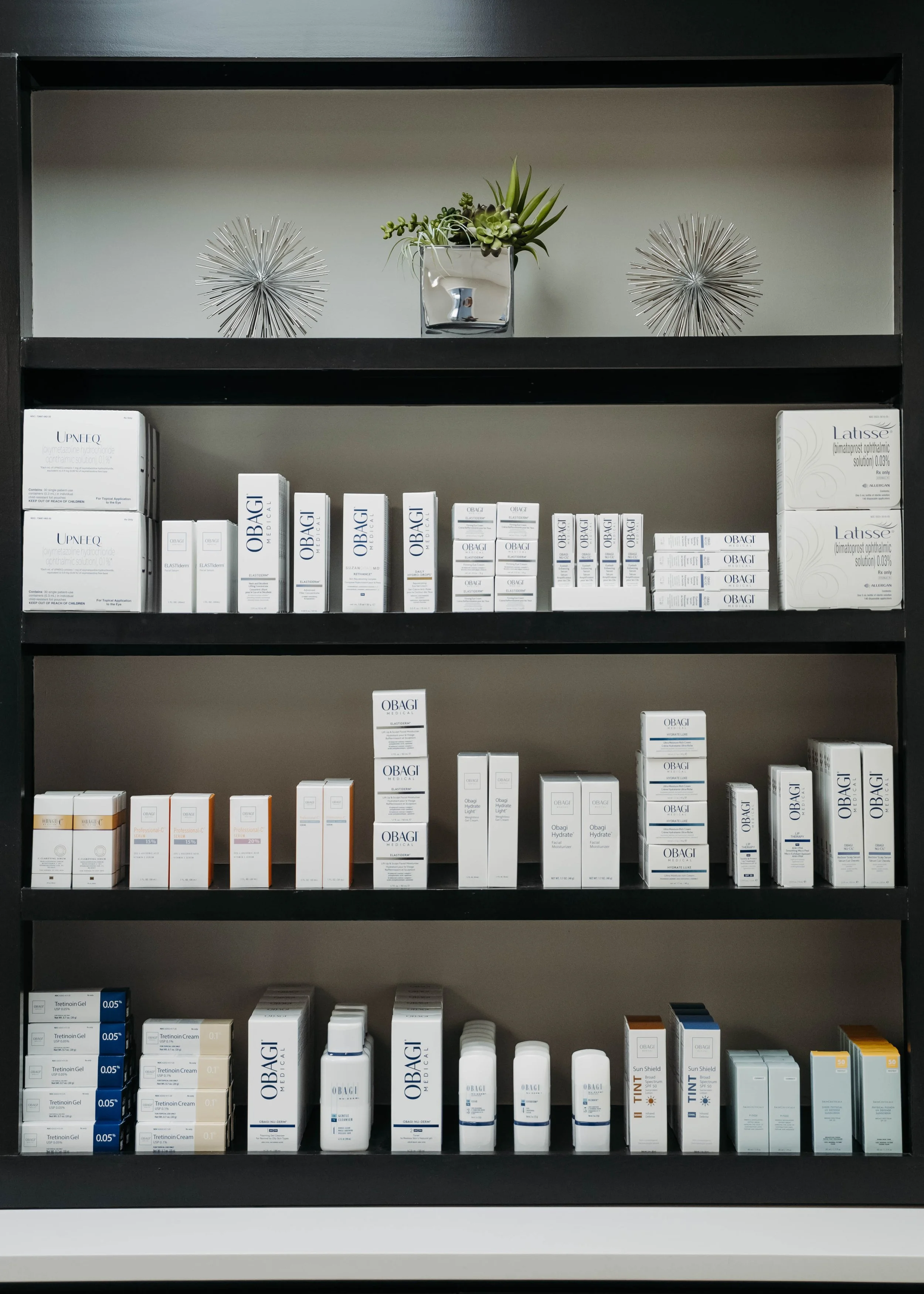 Store shelf with skincare and pharmaceutical products, including OBAGI, UPNEQ, Latisse, Tretinoin gel, and TINT sun protection, topped with decorative metallic sculptures and a small plant.