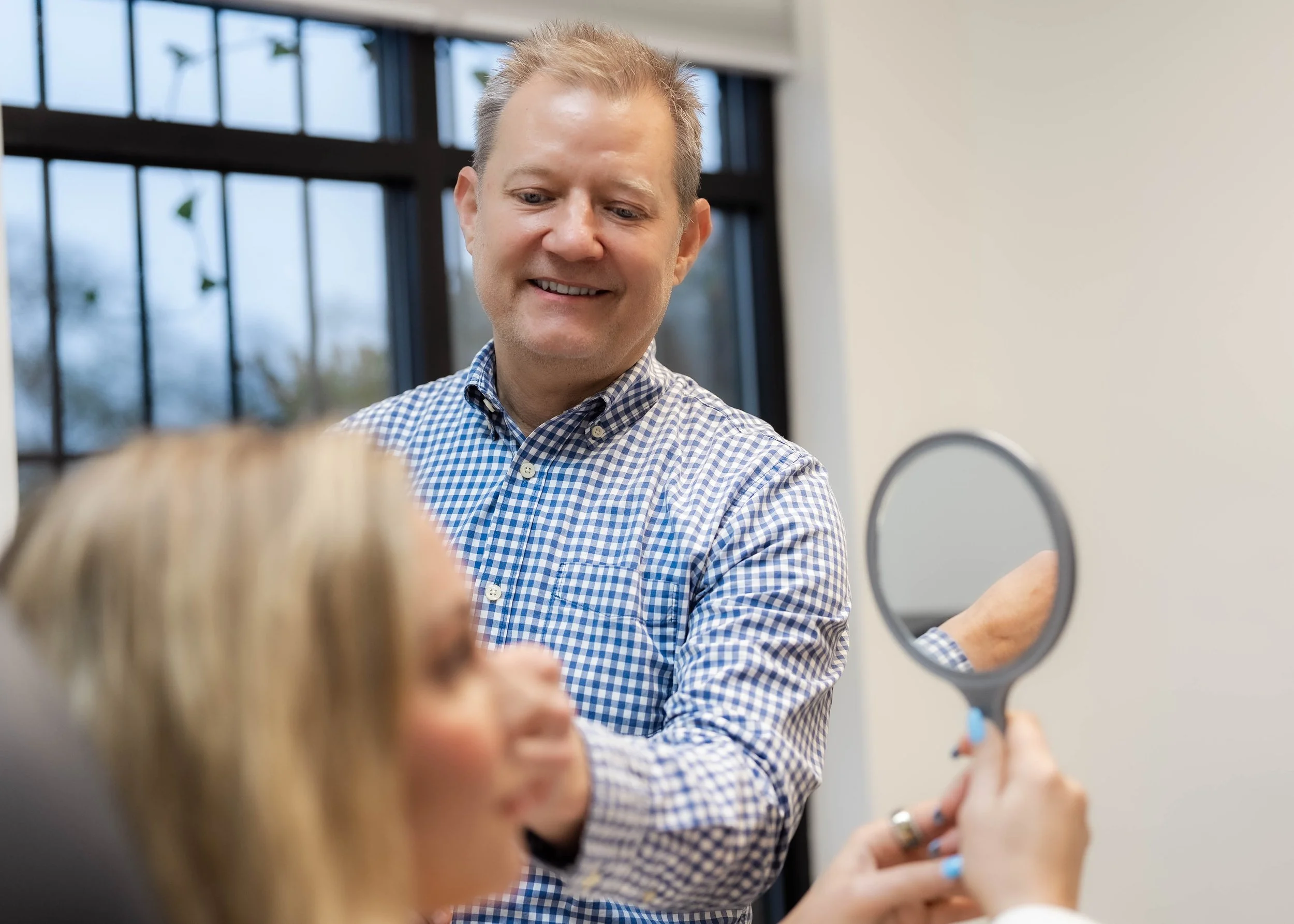 A man in a checkered shirt smiling at a woman holding a small mirror, with a large window in the background.