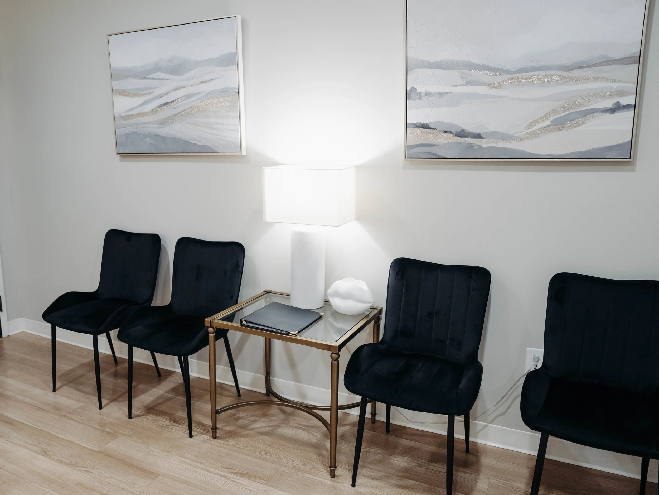 Waiting room with four black chairs, a glass top table with a black notepad, a white lamp, and a decorative white object, with abstract landscape paintings on a white wall.
