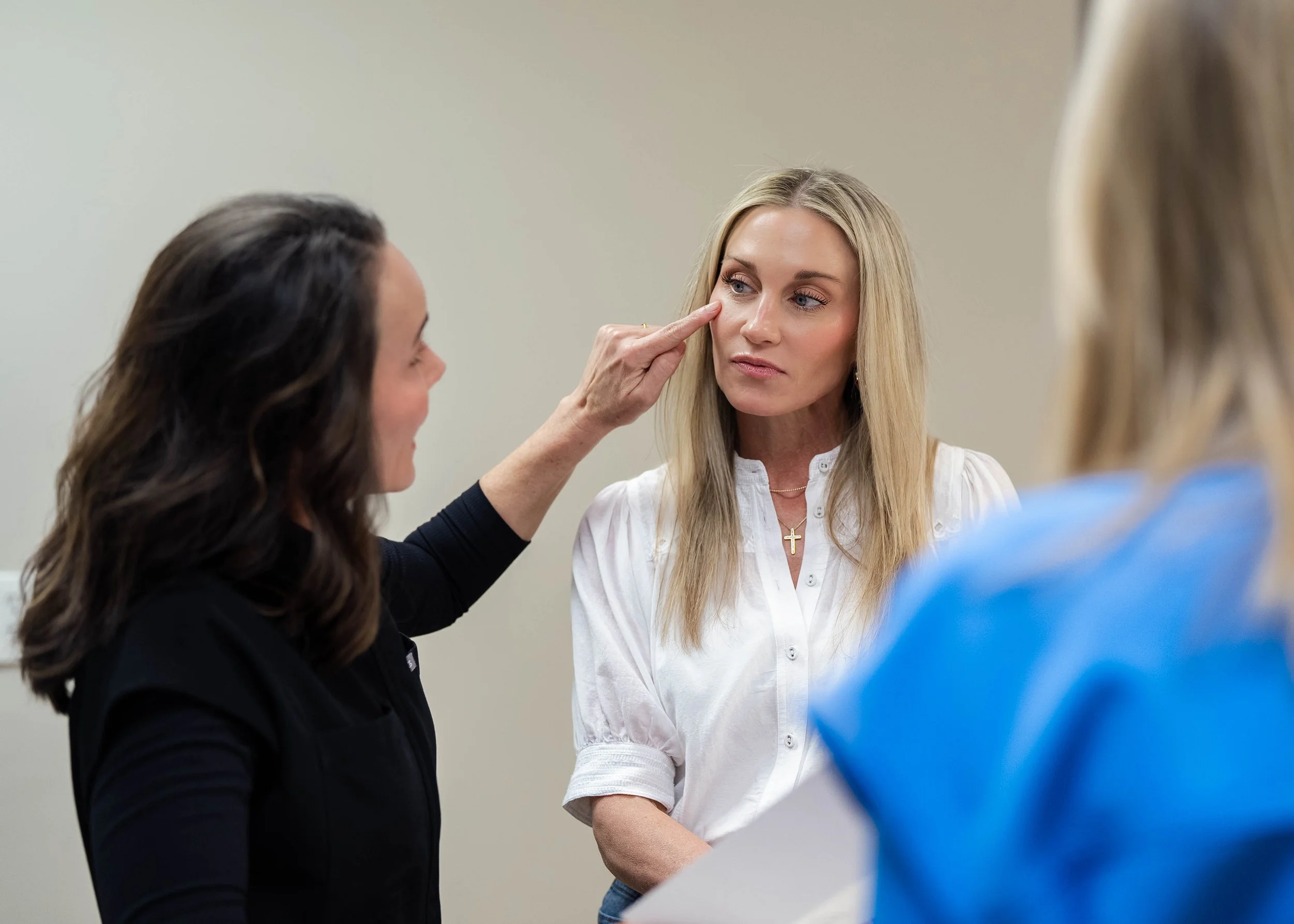 A woman in black applies makeup or skincare to another woman's face, focusing on her nose and cheekbone, while another woman in blue observes in the background.