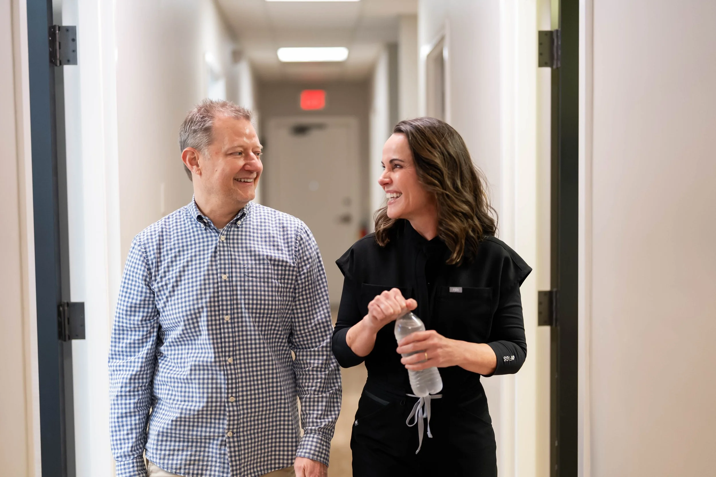 A man and a woman walking and smiling in a hallway, the woman holding a water bottle.