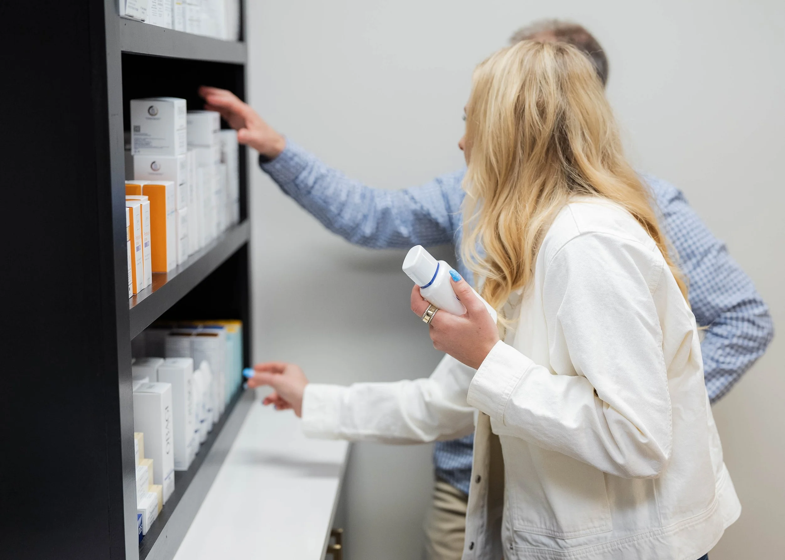 Two people, one woman with long blonde hair and a man with gray hair, are looking at and reaching for medication bottles on a black shelving unit in a pharmacy or medical storage room.