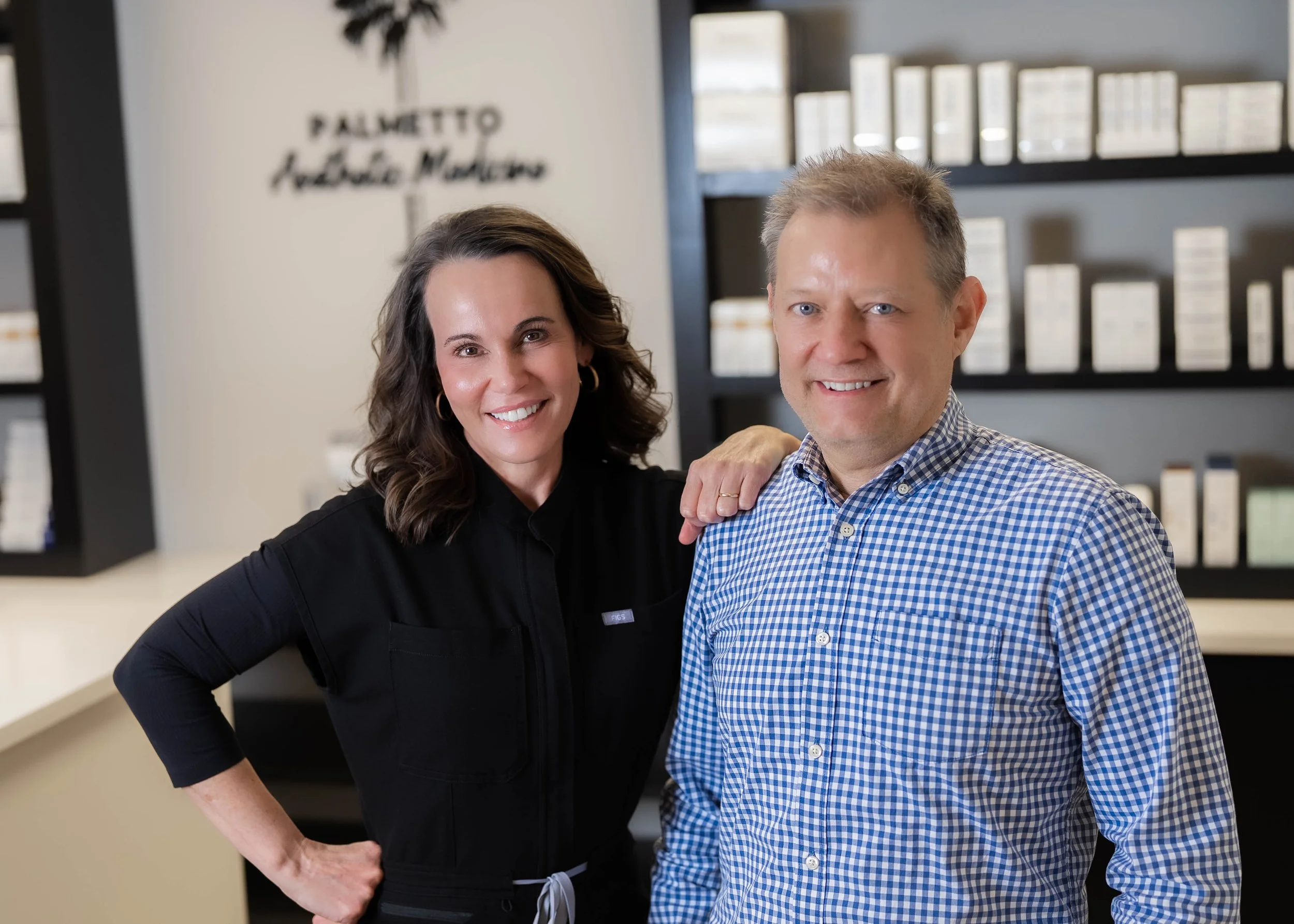 Two smiling people, a woman with shoulder-length brown hair wearing a black top, and a man with short light brown hair in a blue checkered shirt, standing together in an office with shelves of boxed products and a sign reading 'Palmetto' in the background.