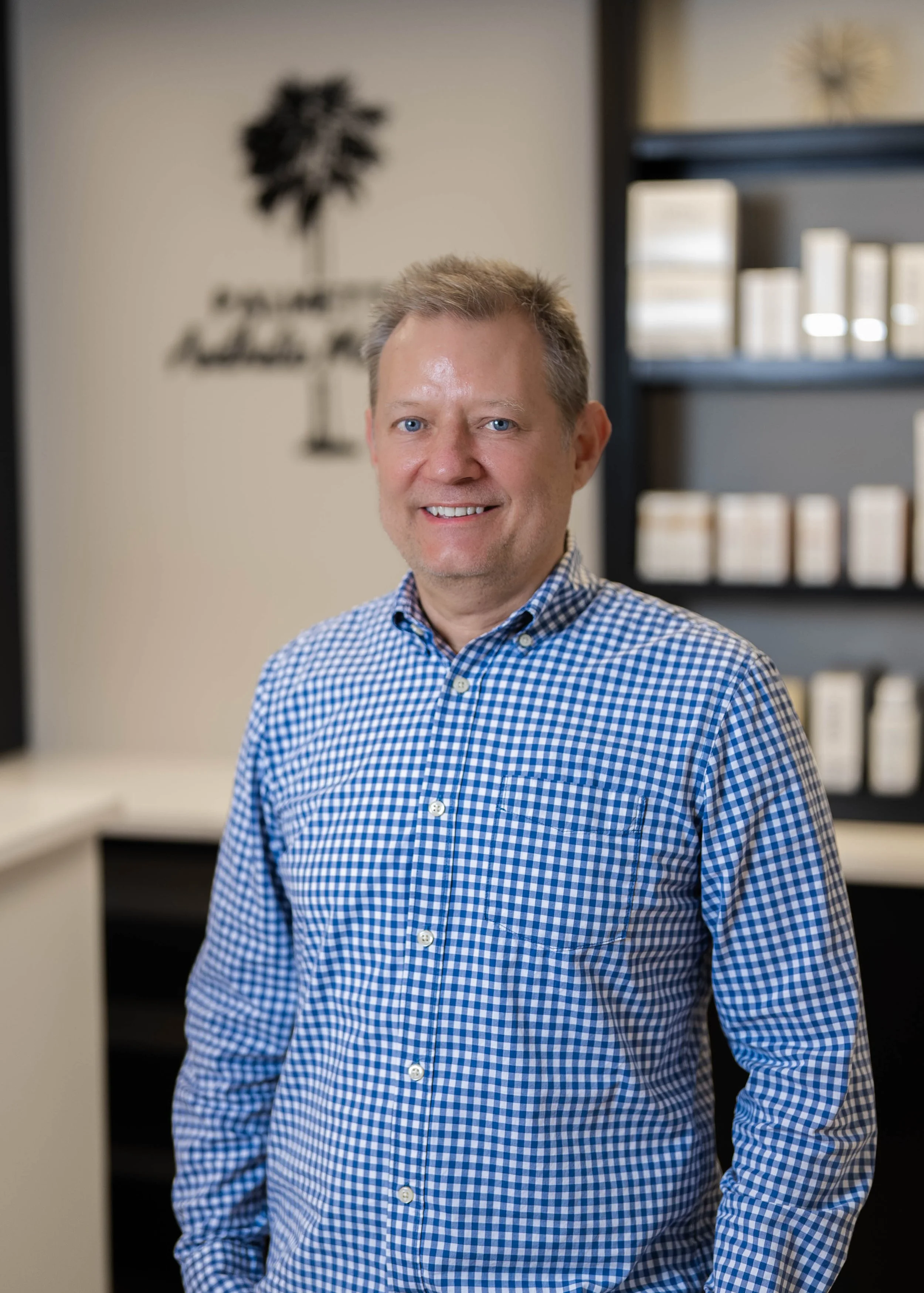 A man with short blond hair and blue eyes, wearing a blue and white checkered shirt, smiling, standing in an indoor office space with shelves and a wall decoration in the background.