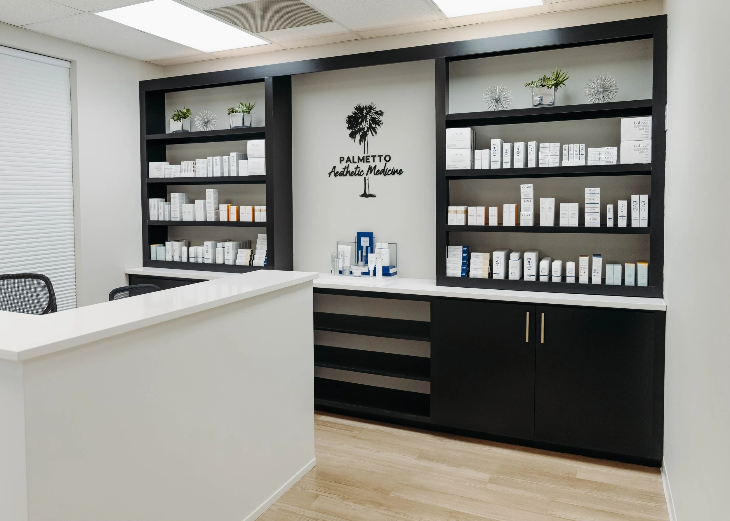 Interior of a medical aesthetic clinic with black shelving units filled with skincare products, a white reception desk, and the clinic's logo on the wall reading 'Palmetto Aesthetic Medicine' with a palm tree graphic.