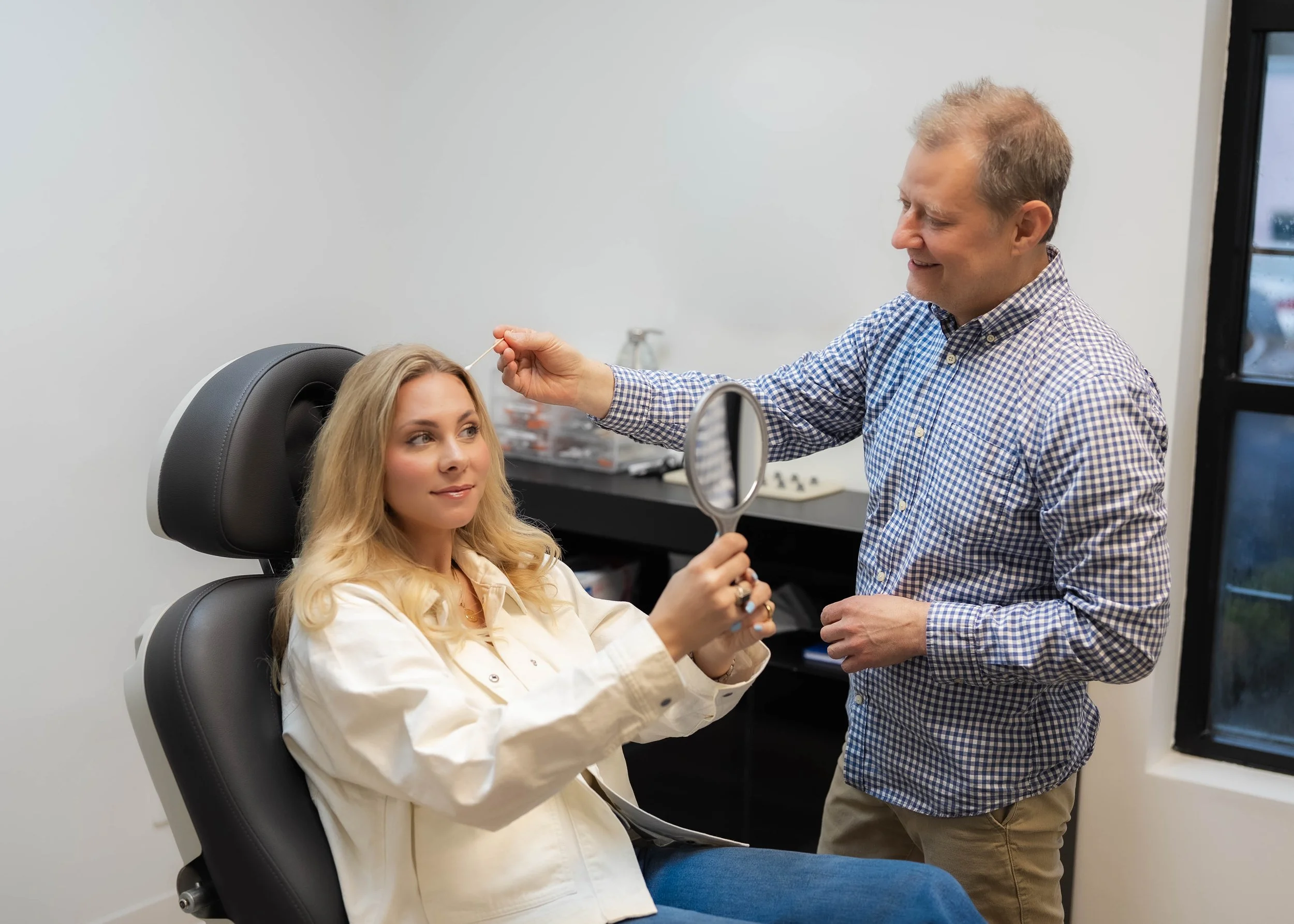 A woman is sitting in a black medical chair at a clinic while a male healthcare provider is preparing to give her an injection, smiling.