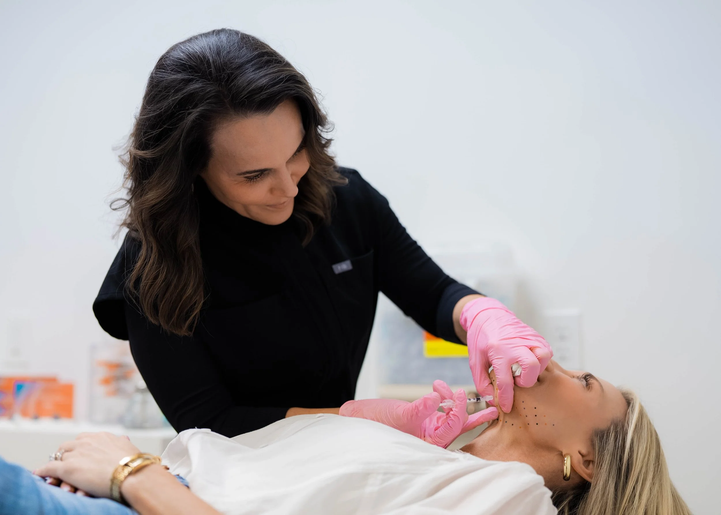 A woman lying on a hospital bed receiving a cosmetic injection in her cheek from a healthcare professional wearing pink gloves.