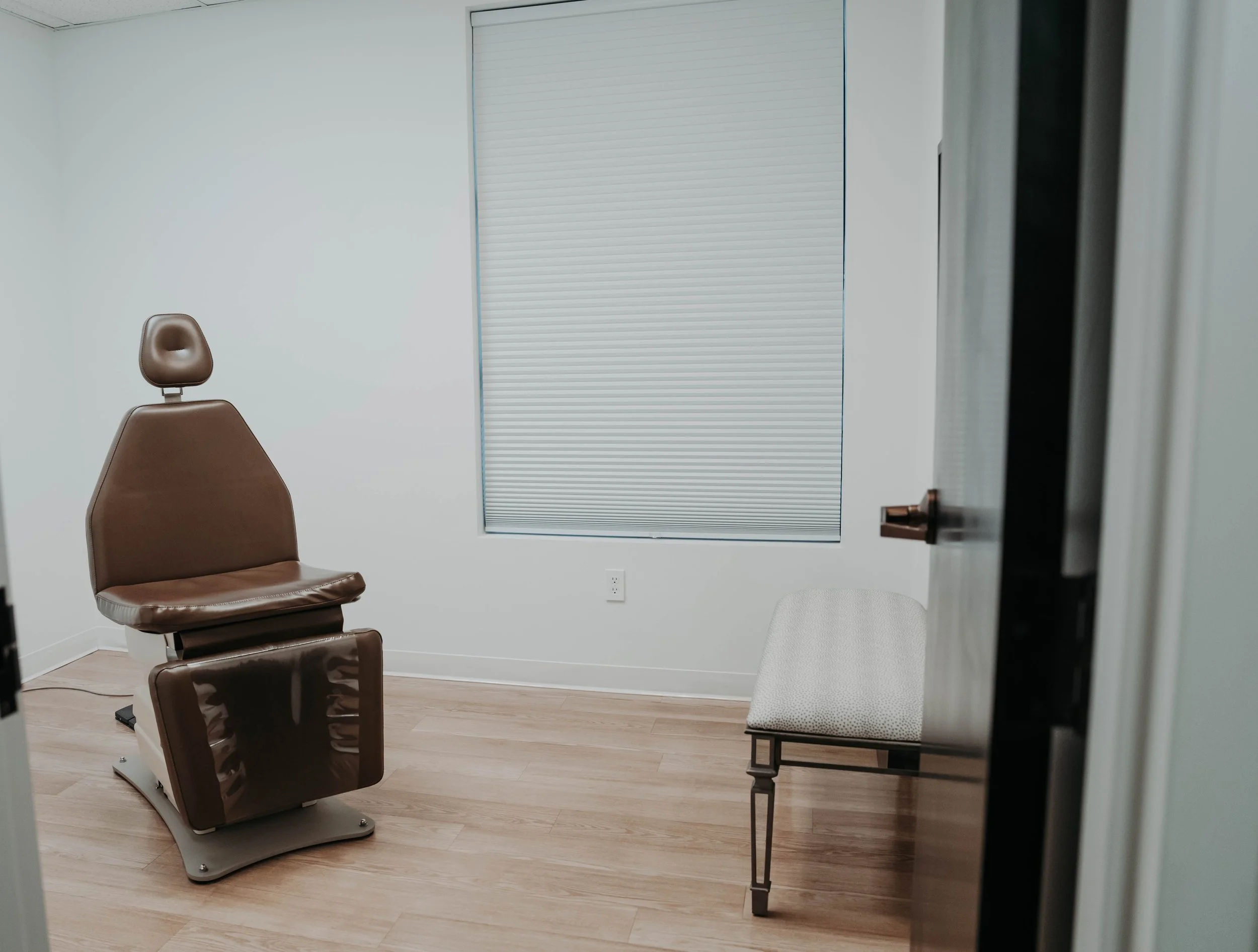 A medical examination room with a brown examination chair, a small white cushioned stool, a window with white blinds, and a partially open door.