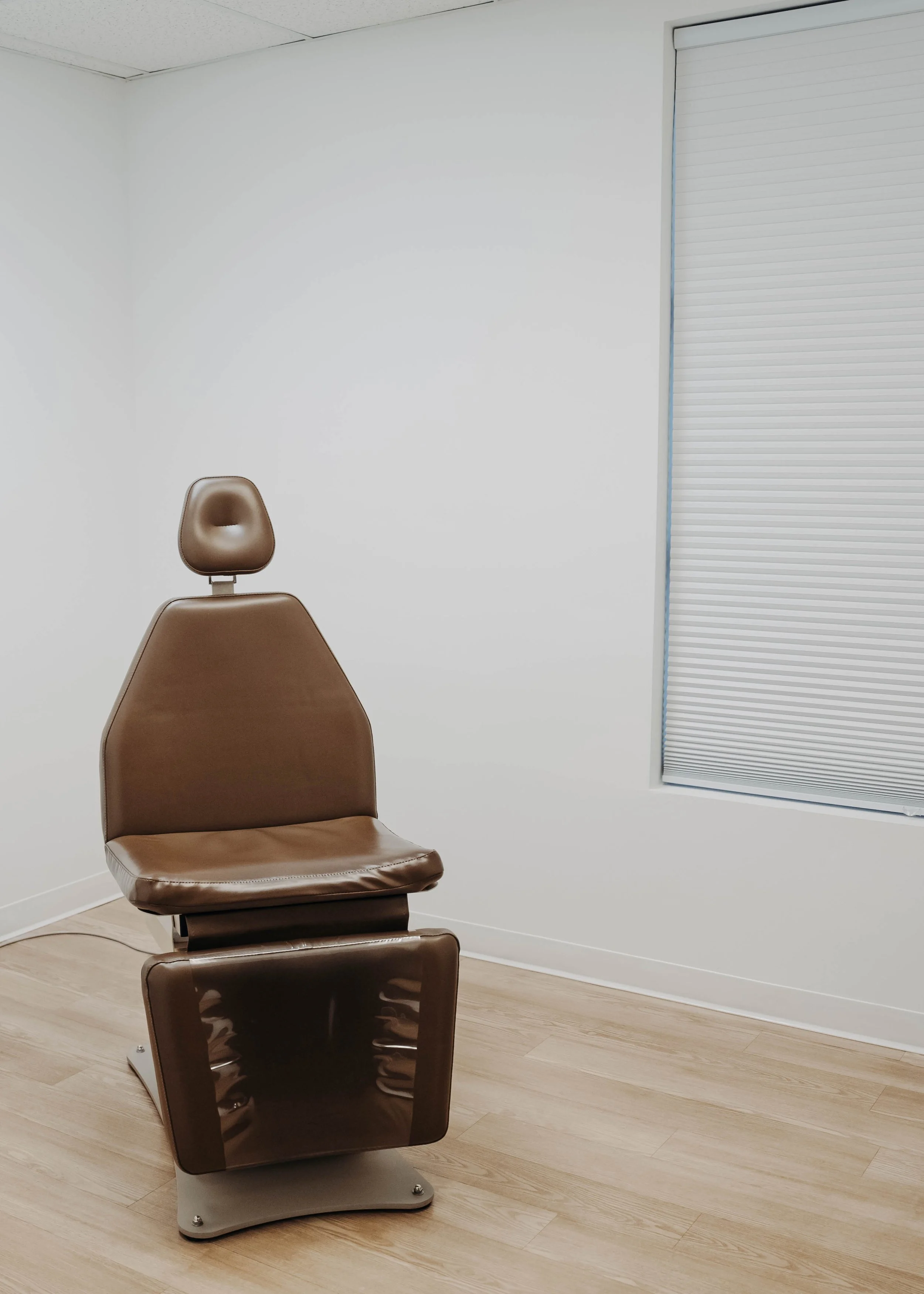 A brown examination chair with a headrest in a medical or clinic room with plain white walls, a window with closed blinds, and light-colored wooden flooring.