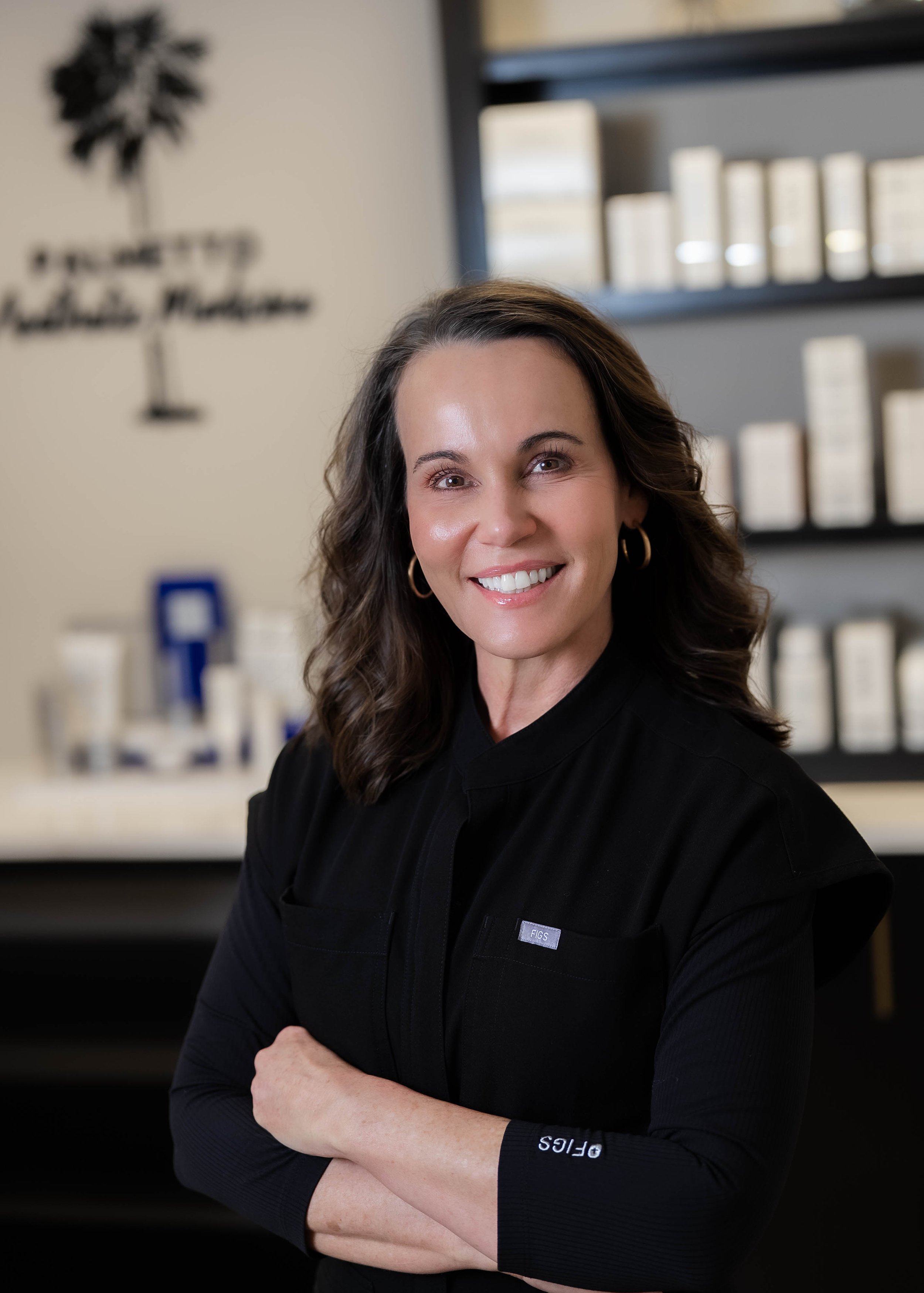Smiling woman with brown wavy hair, wearing black clothing with a small 'FIGS' logo on sleeve, standing in a room with shelves and a wall art logo in the background.